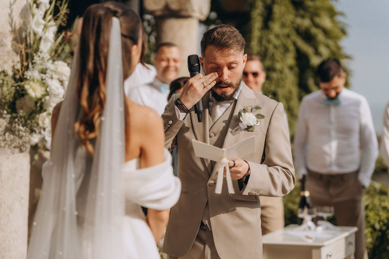 Emotional groom wiping tears during outdoor wedding ceremony while bride stands before him in white gown.