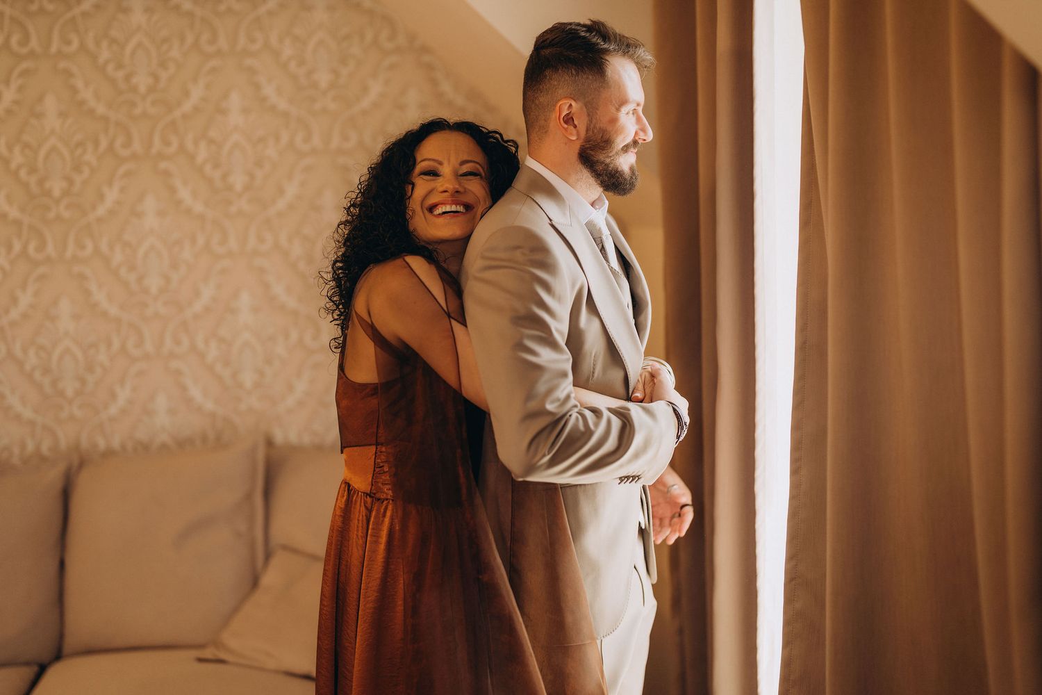 Smiling bride in rust dress embraces groom in gray suit near window in elegant wedding preparation room.