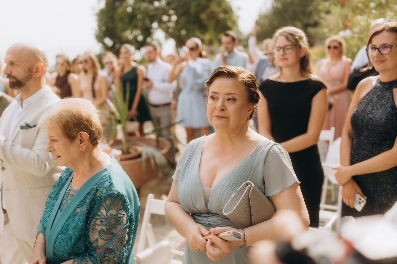 Guests in elegant attire attend an outdoor wedding ceremony on a sunny day.