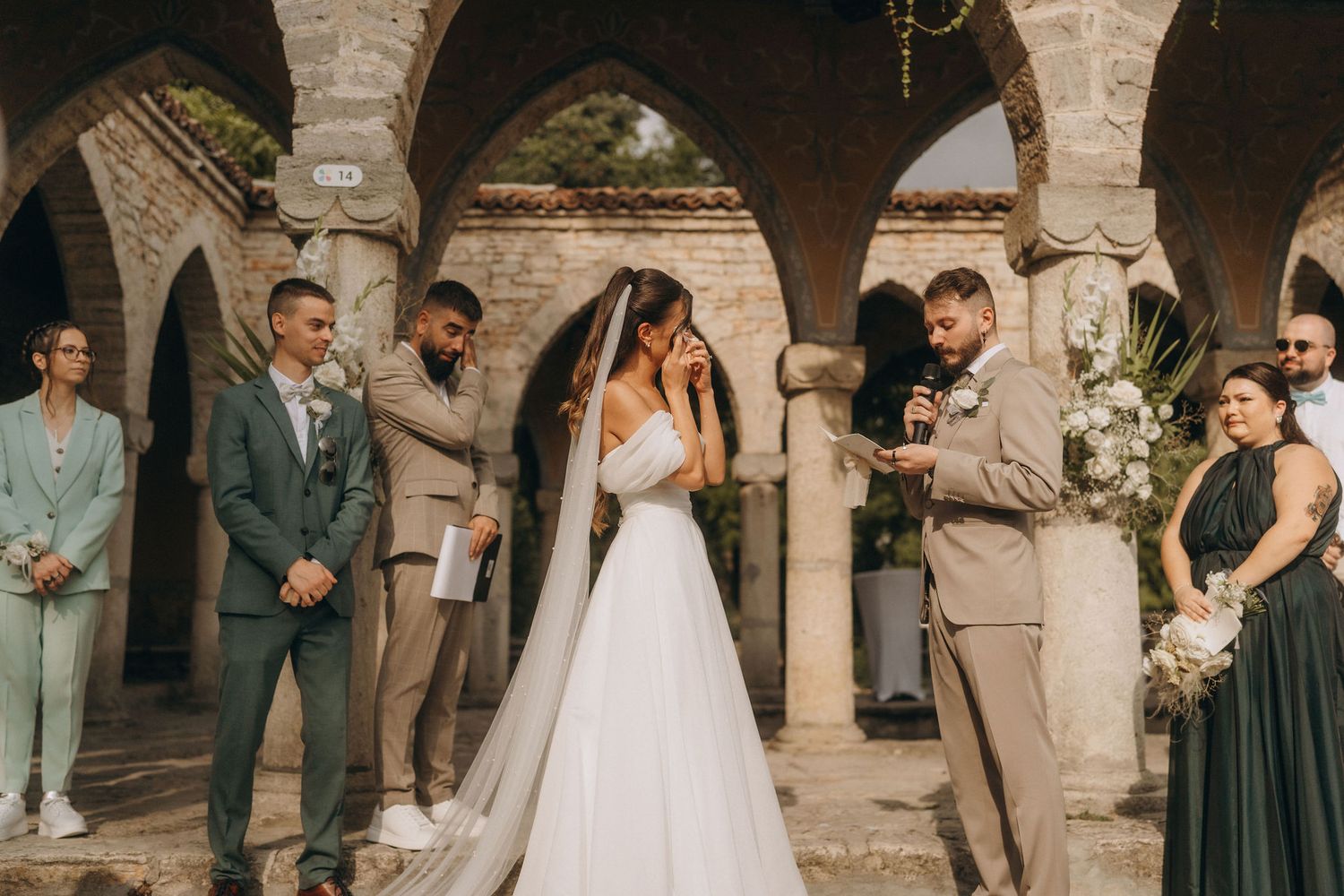 Outdoor wedding ceremony at rustic stone arches with bride in white gown, groom in tan suit, and wedding party in green attire.