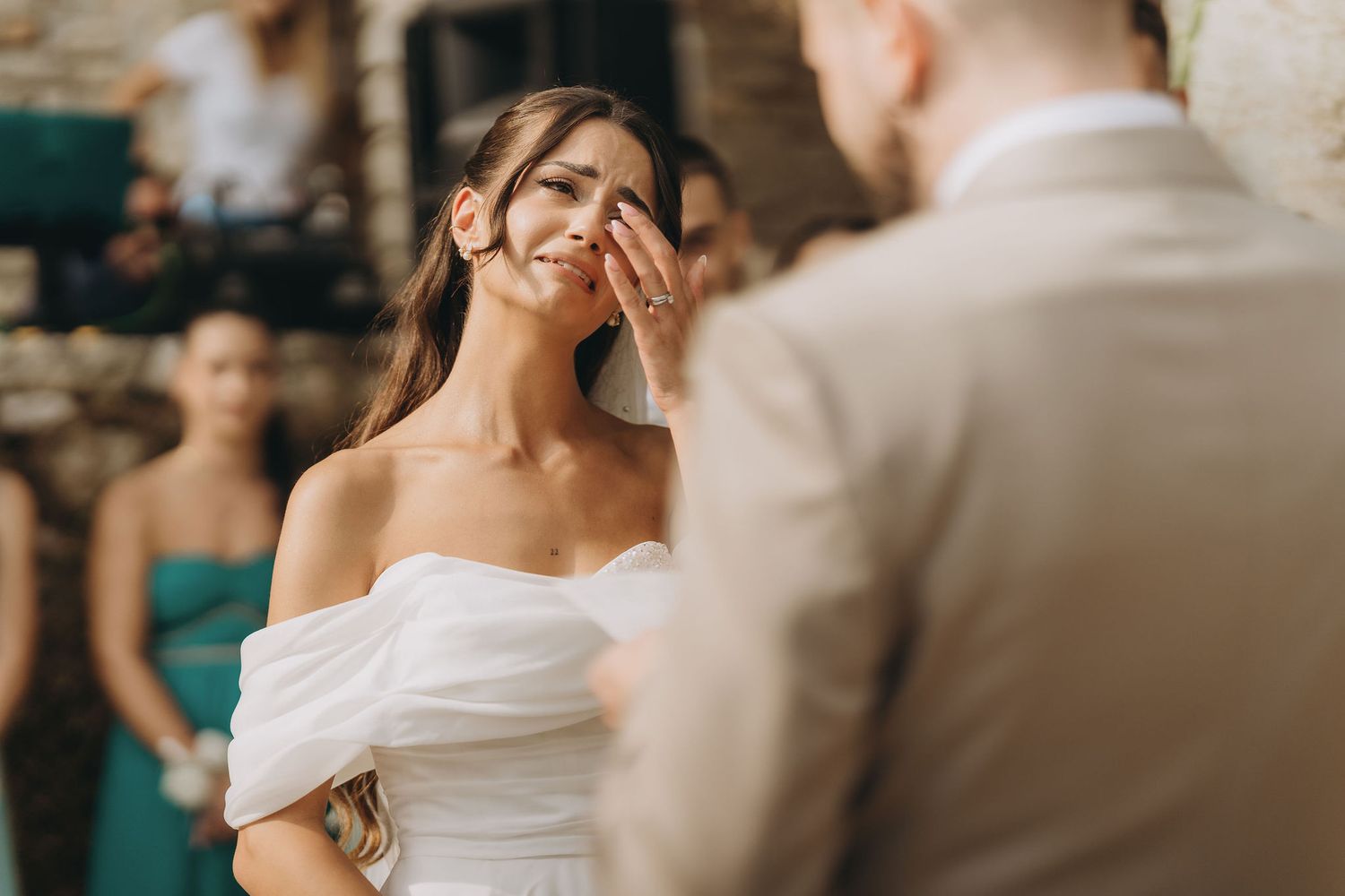 Groom gently wipes tears from smiling bride's cheek during emotional outdoor wedding ceremony.
