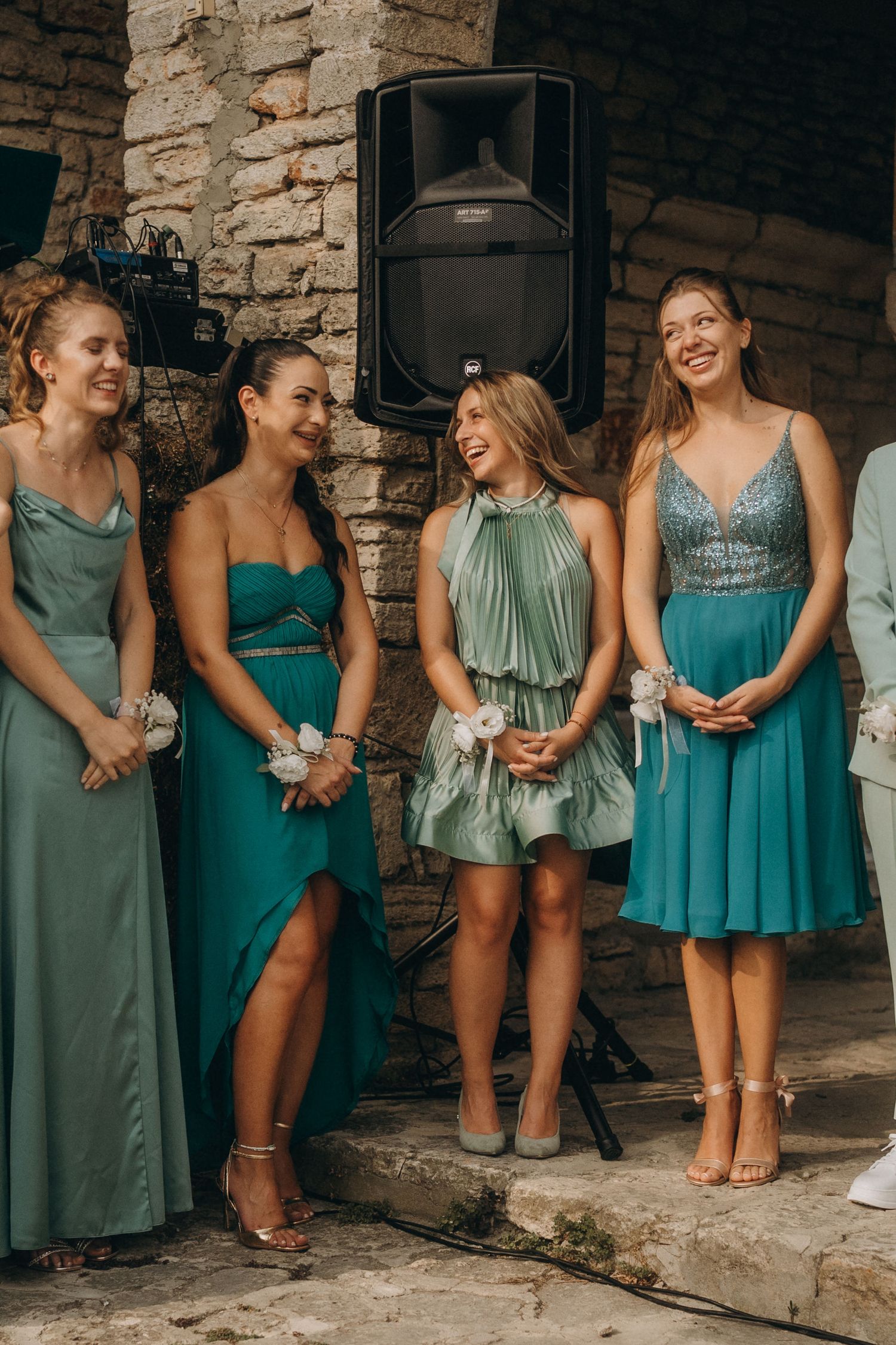 Four bridesmaids in elegant teal and green dresses laughing together at an outdoor wedding reception.