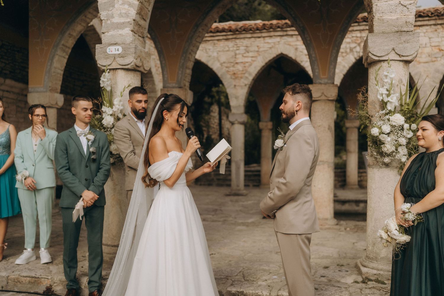 Bride reads vows to groom during outdoor wedding ceremony at rustic stone venue with arched columns.