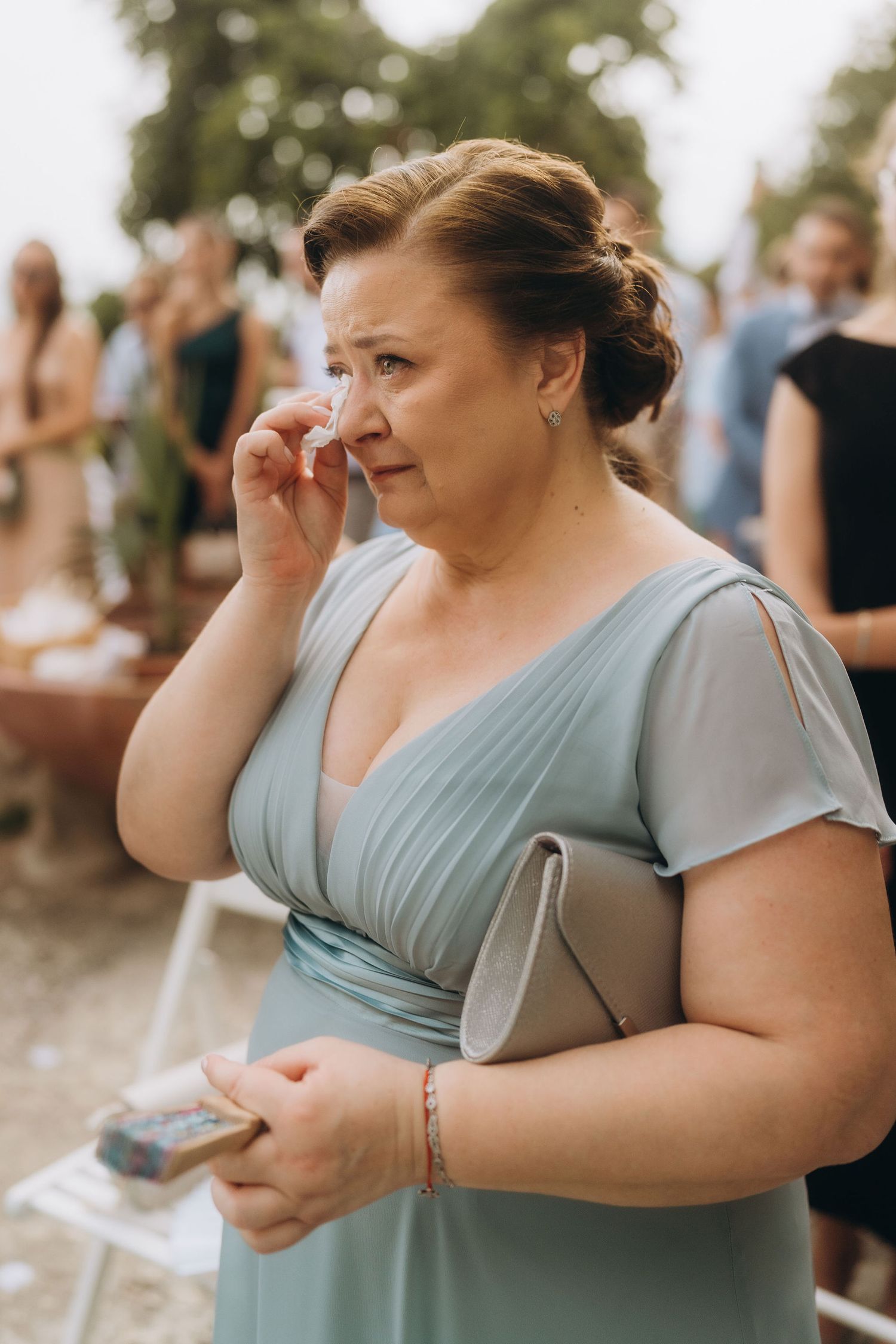 Emotional woman in blue dress wiping tears at outdoor wedding ceremony, holding clutch purse.