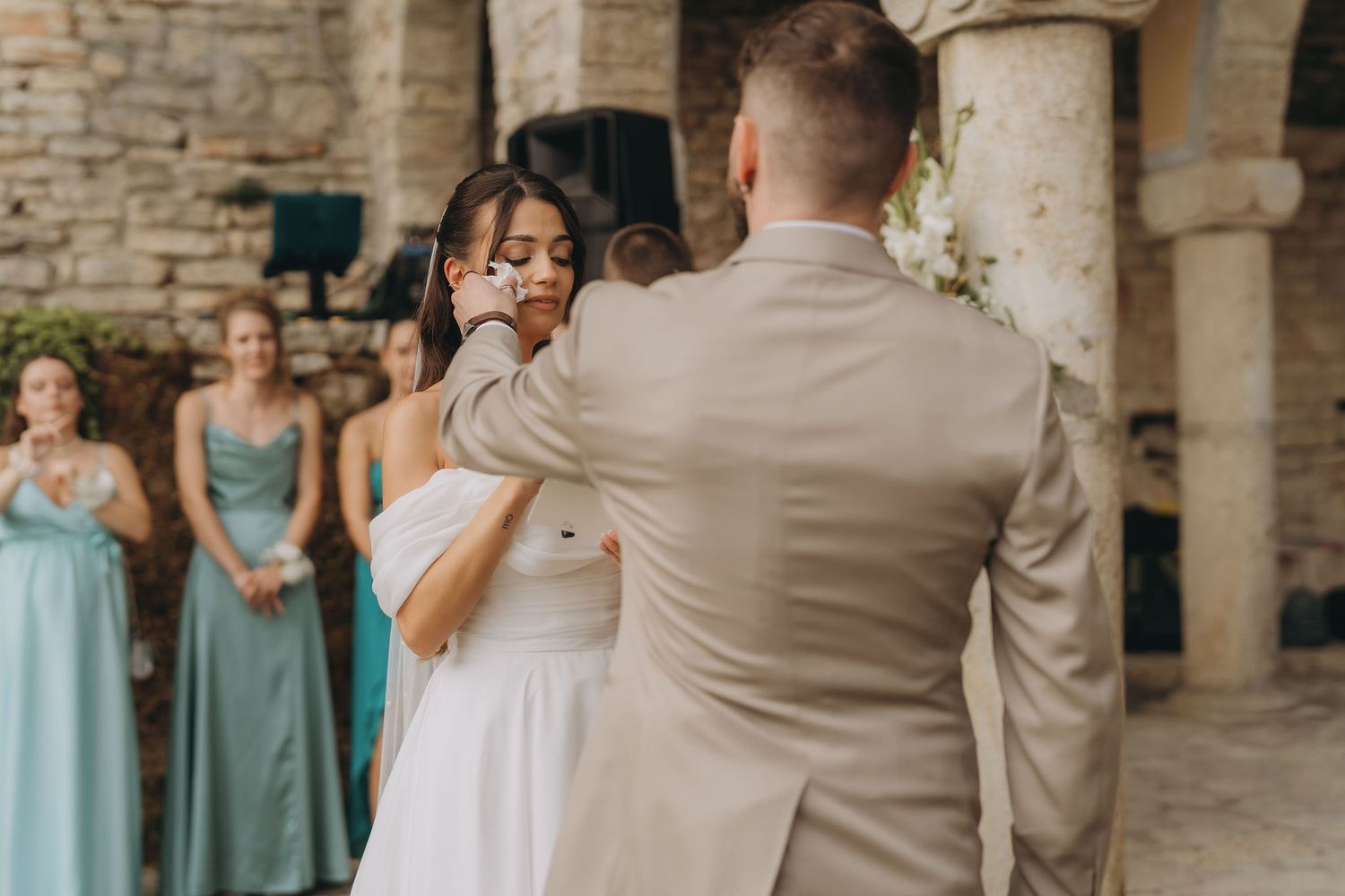 Groom tenderly wiping tears from brides cheek during emotional first dance at outdoor stone venue wedding.