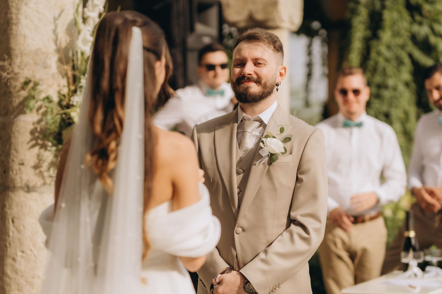 Groom in beige suit smiling at bride during outdoor wedding ceremony with guests in background.