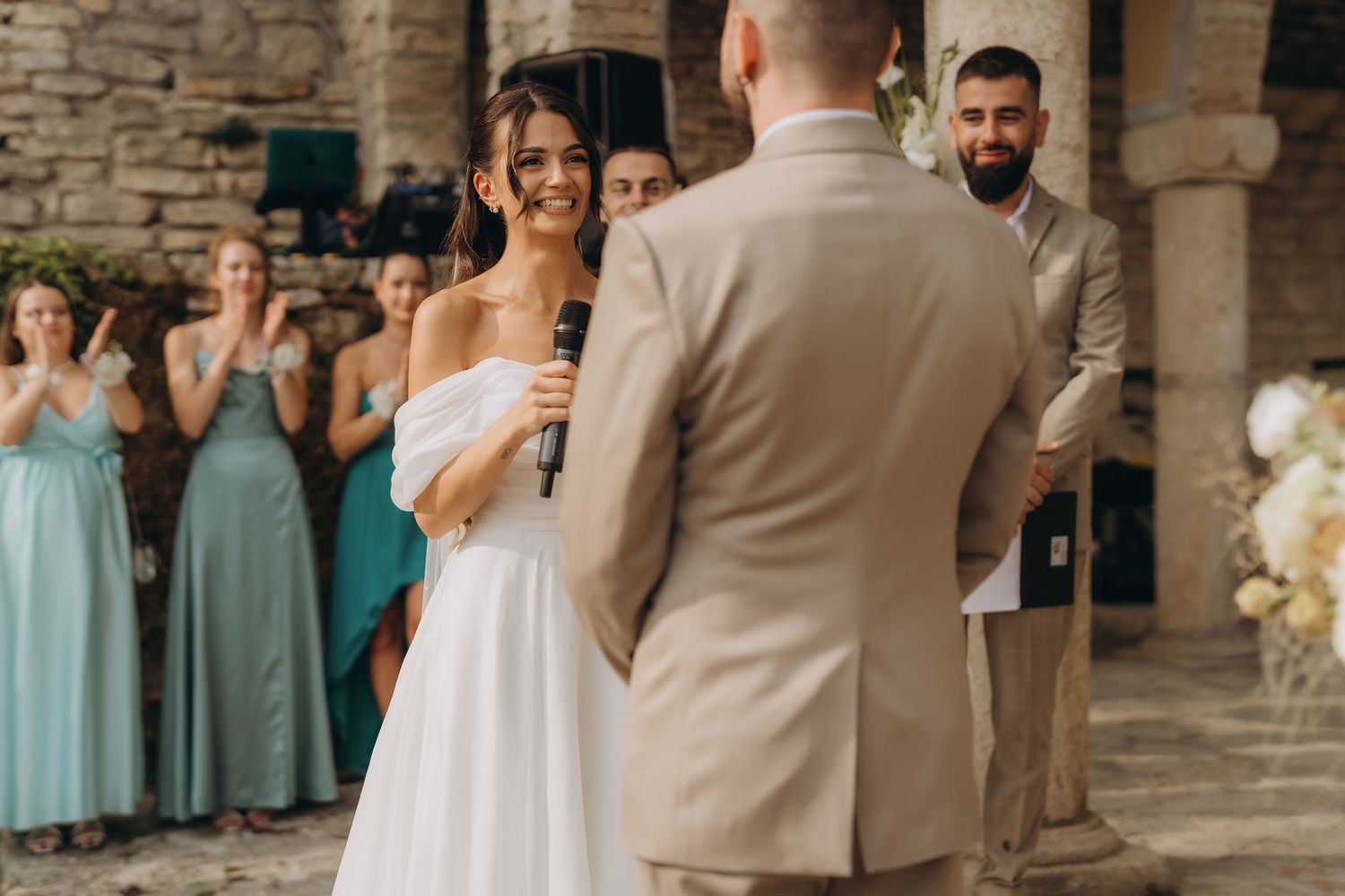Bride smiles at groom during outdoor wedding ceremony with guests in teal dresses in background.