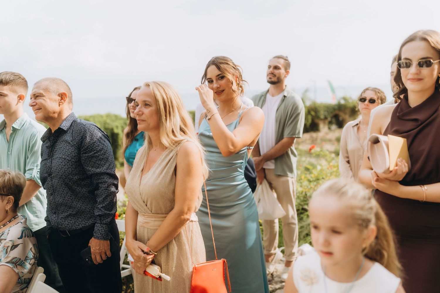 Elegantly dressed wedding guests mingling outdoors on a sunny day, women in beige and teal gowns wearing sunglasses.