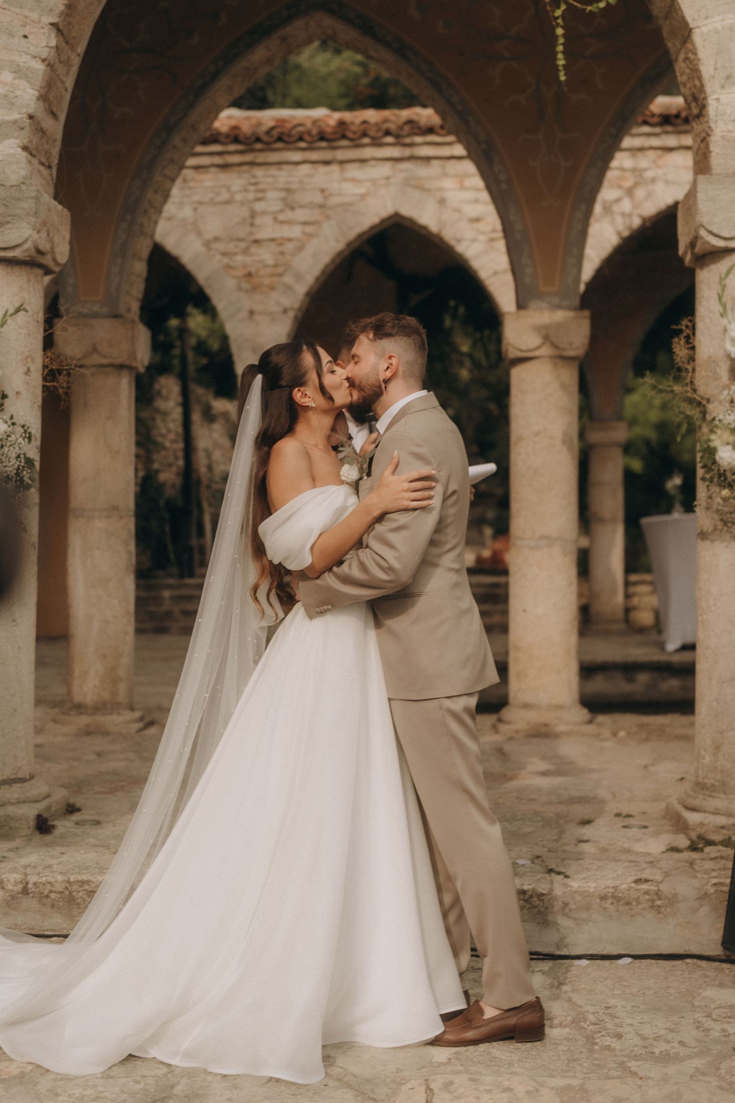 Bride and groom share a romantic kiss in a white gown and tan suit under stone arches at a rustic wedding venue.