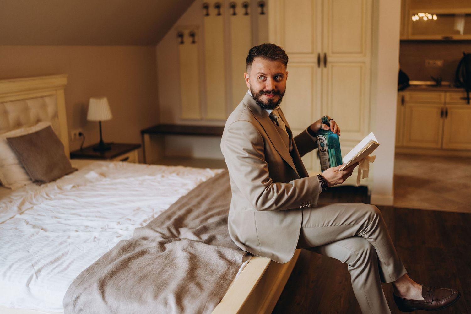Bearded man in beige suit sitting on bed holding book and bag, smiling in elegant hotel room.