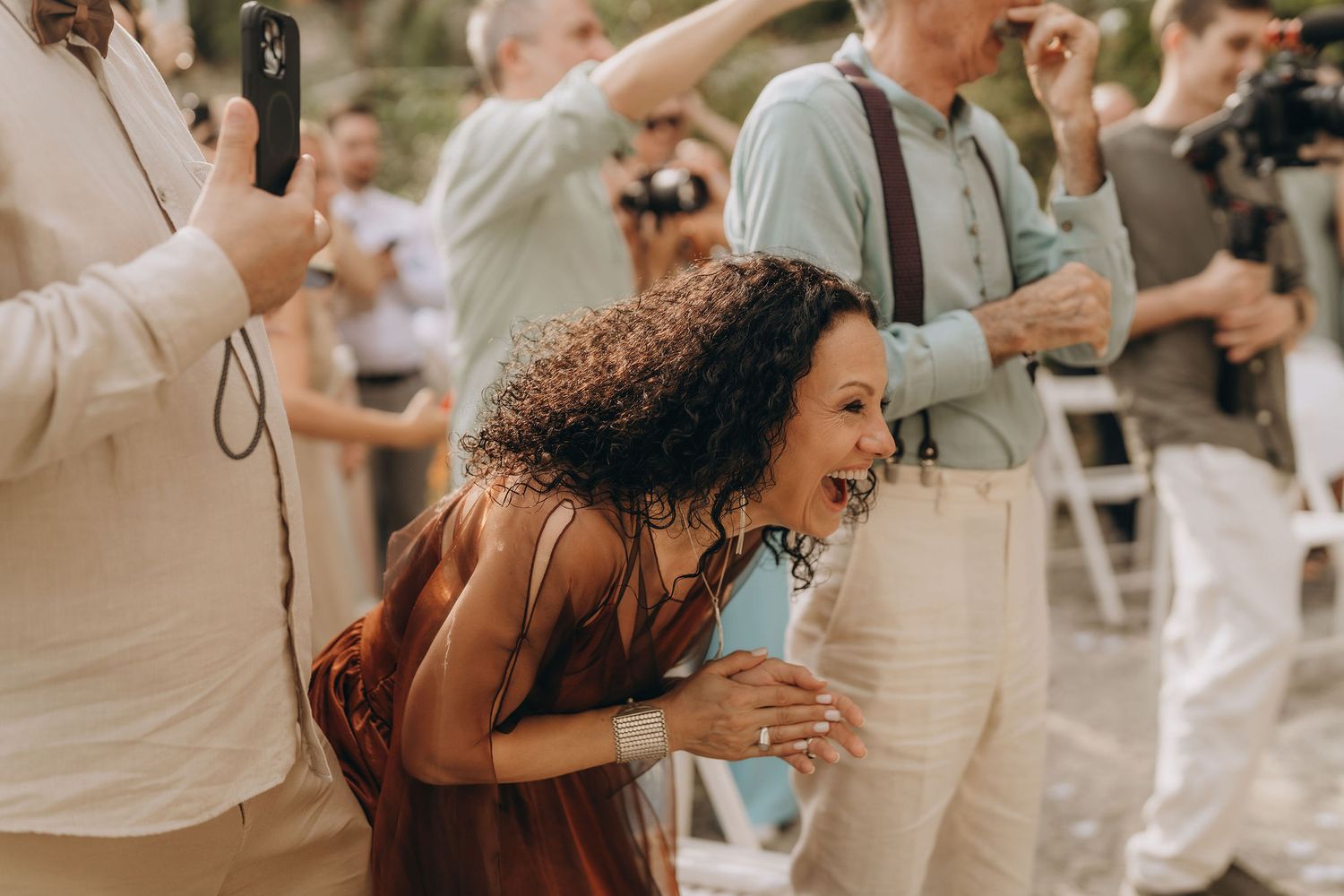 A joyful woman with curly hair laughs and bends forward at an outdoor wedding celebration.