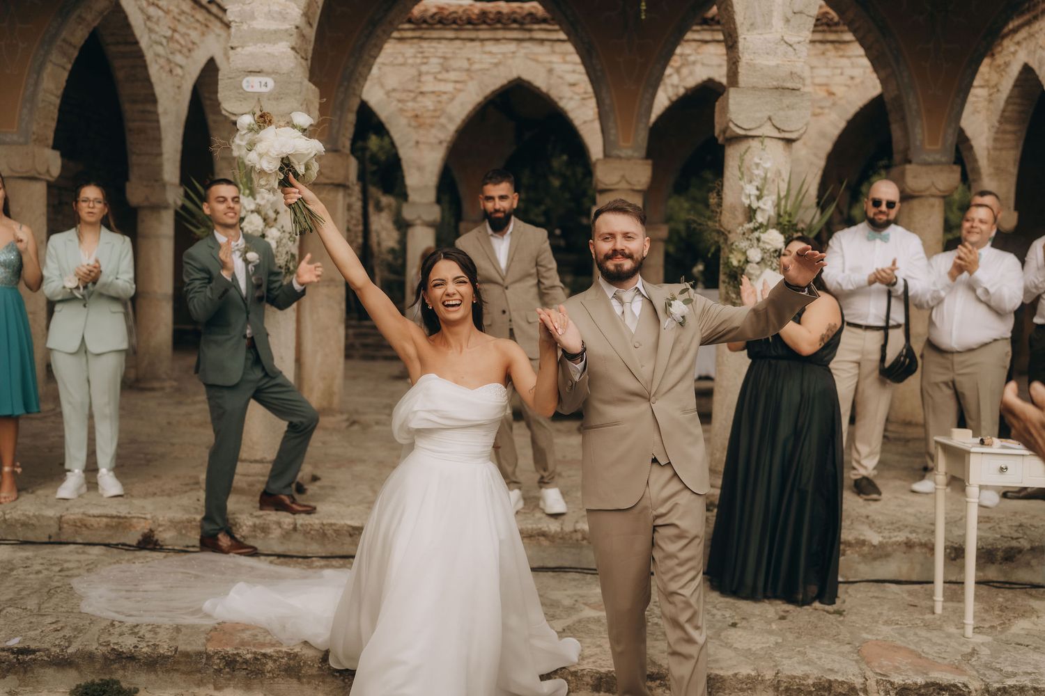 Joyful newlyweds celebrate their wedding ceremony in a historic stone courtyard, surrounded by applauding guests.