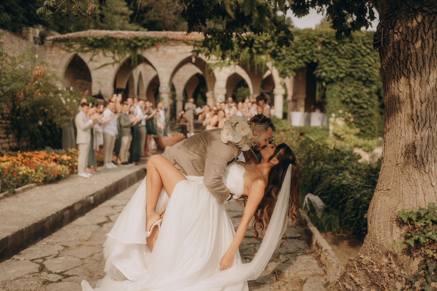 Groom dips bride in romantic kiss at outdoor garden wedding with guests celebrating in background.