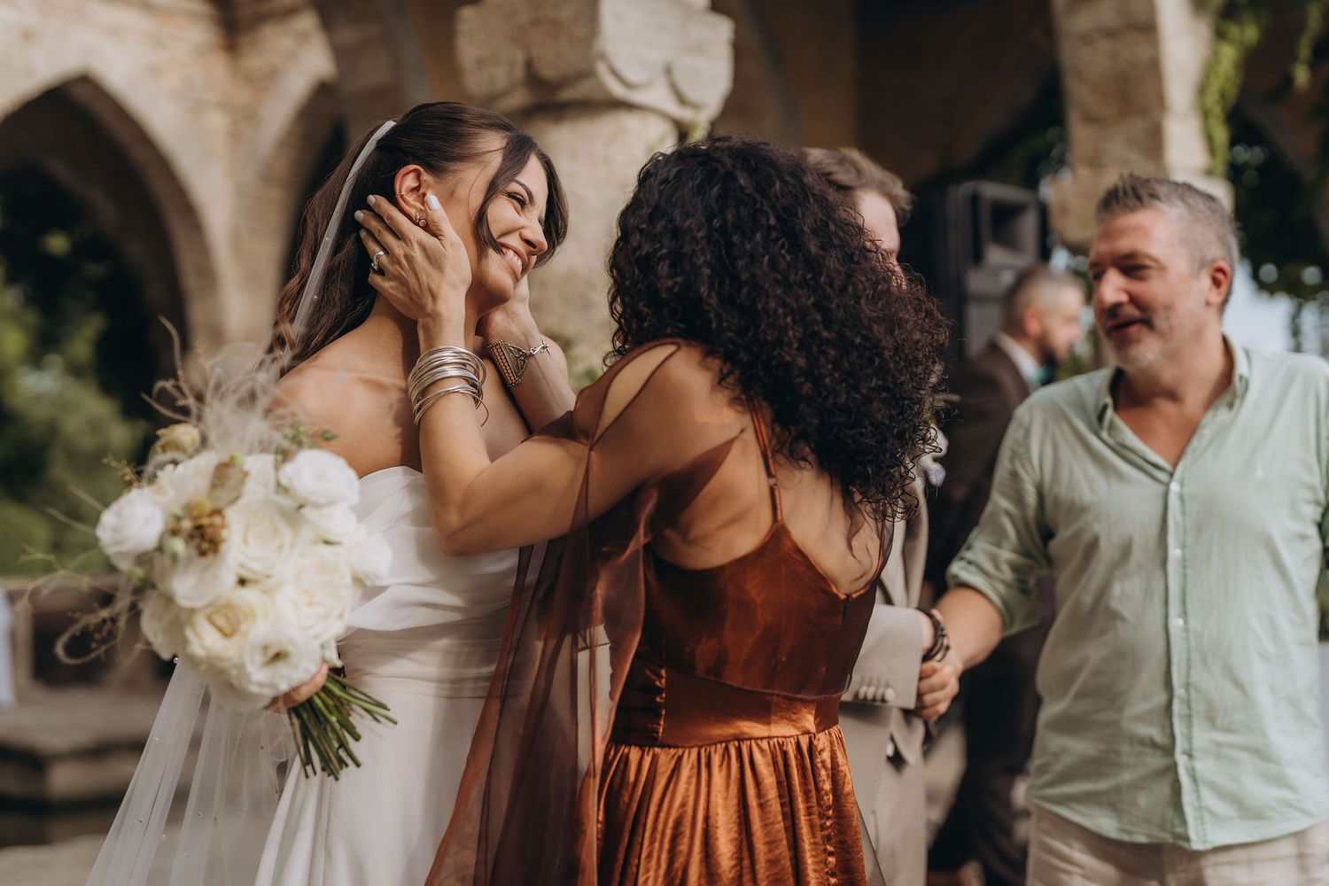 Bride in white gown holding bouquet embraced by guest in rust dress at outdoor wedding celebration.