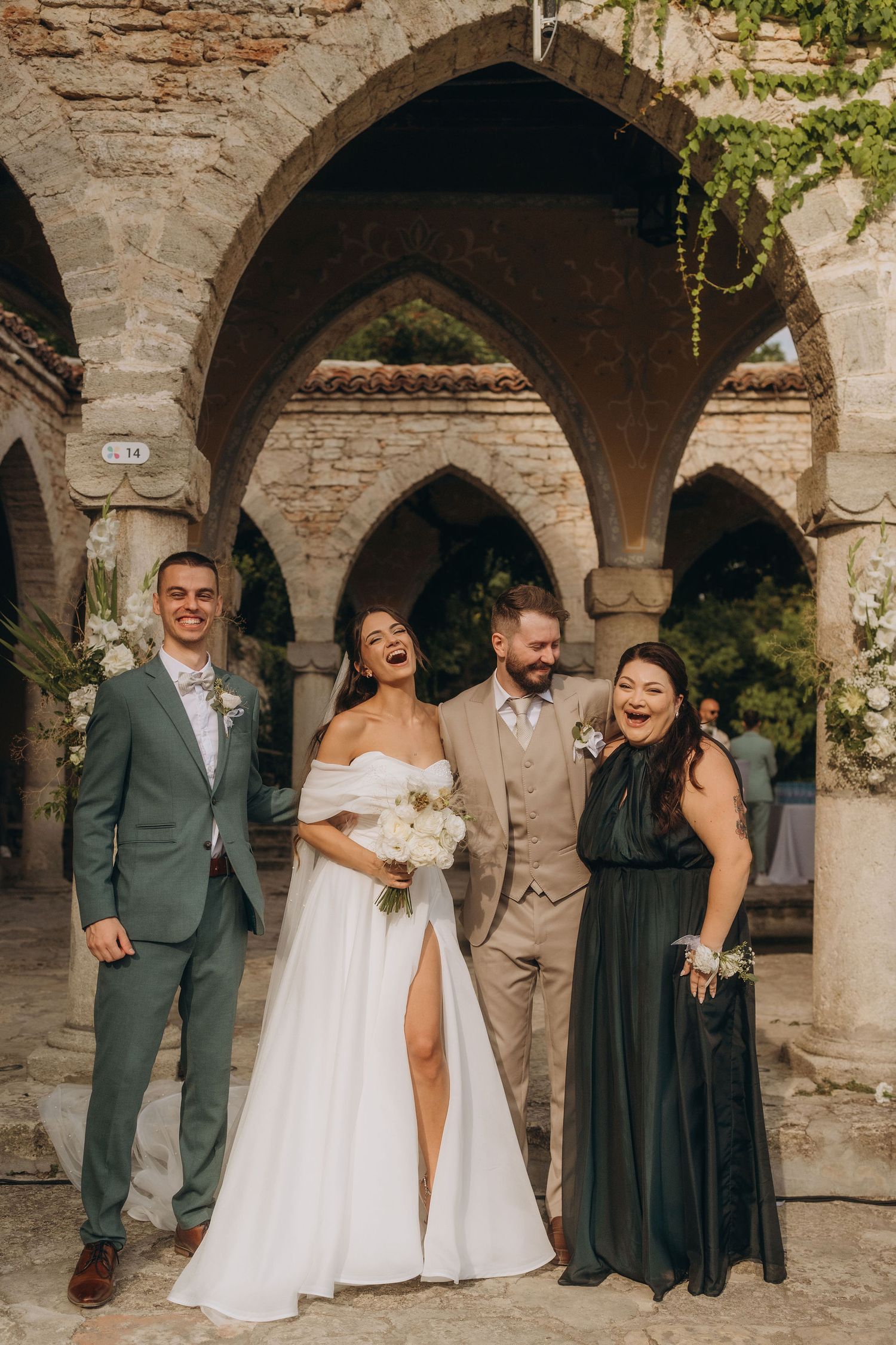 Bride and groom laugh with wedding guests under stone arches at an outdoor venue.