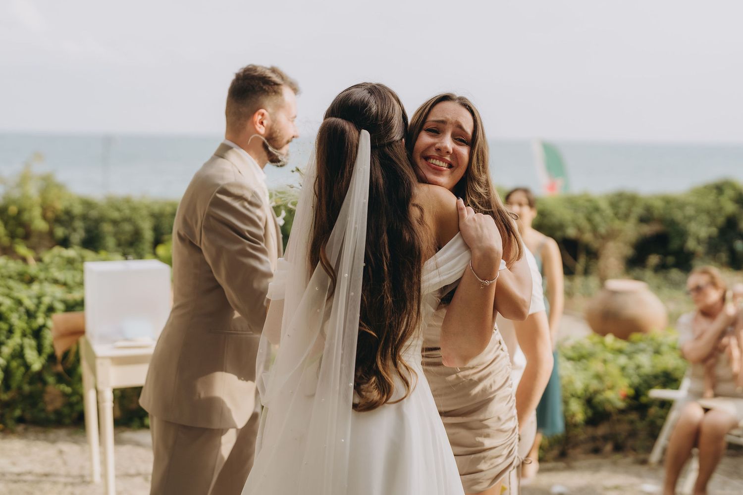 Bride hugging smiling bridesmaid in gold dress at outdoor wedding ceremony with ocean view.