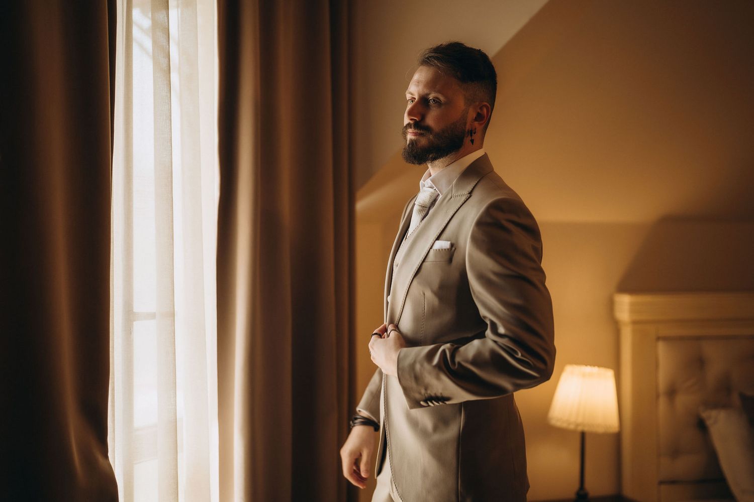 Stylish bearded man in tailored beige suit standing by window in warmly lit hotel room.