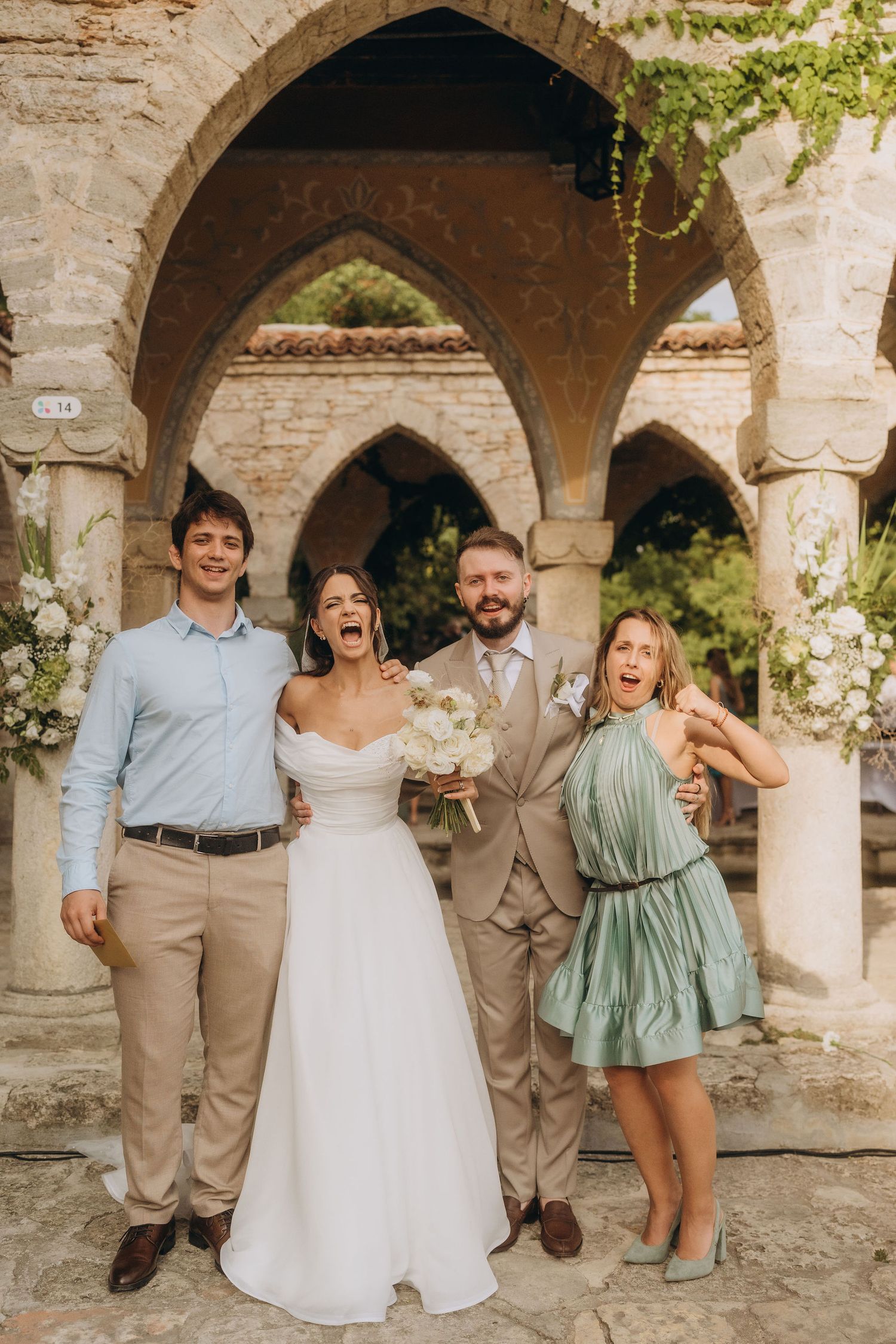 Bride and groom celebrate with two guests under stone arches at an outdoor wedding venue.