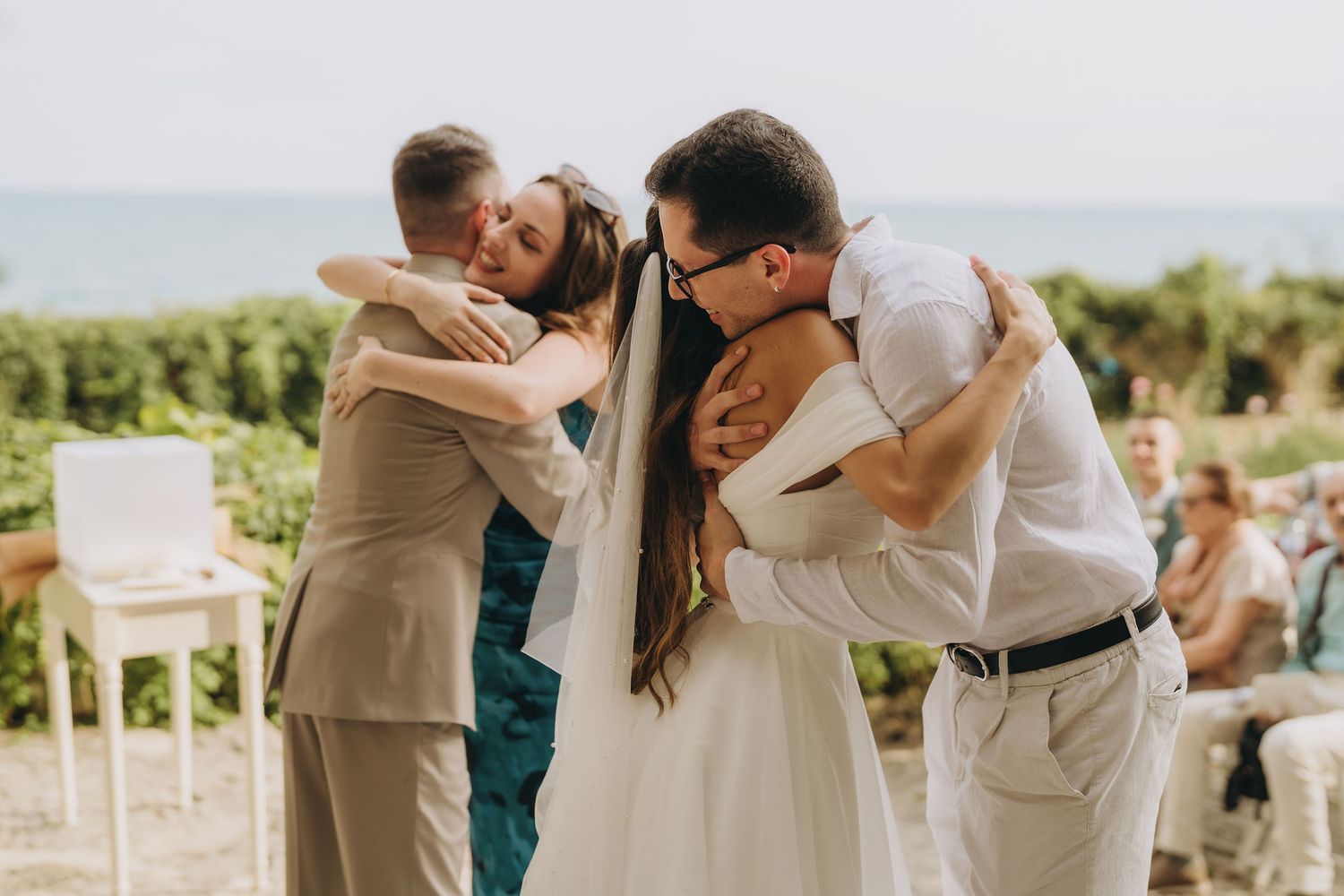 Bride sharing group hug with two men in suits at outdoor seaside wedding ceremony.