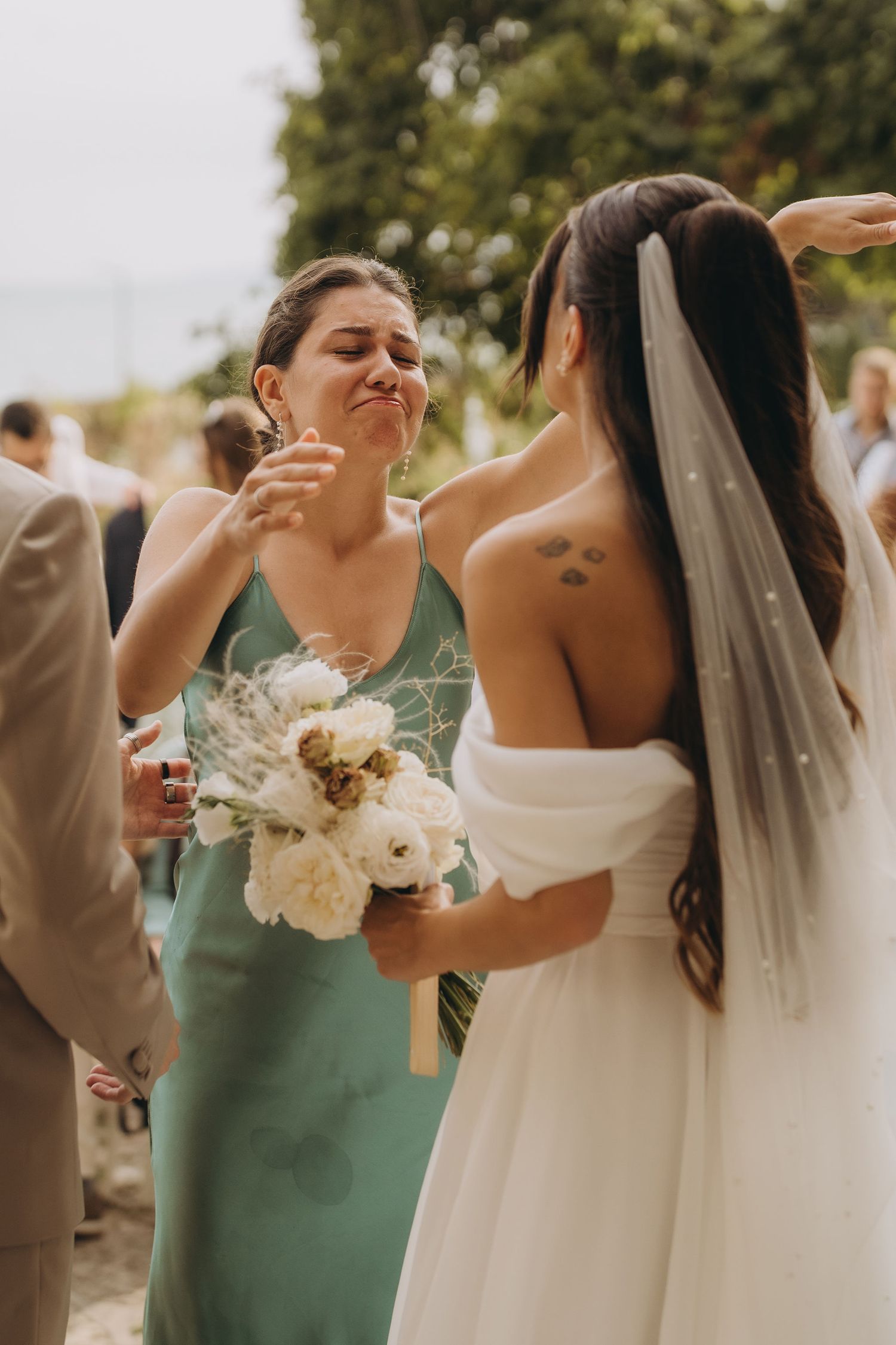 Emotional bridesmaid in green dress holding white bouquet embracing bride in white wedding gown.