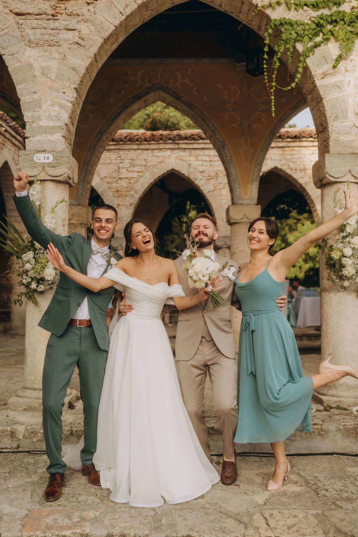 Joyful wedding party celebrating under stone arches, bride in white gown with bouquet, groom and guests in sage green attire.