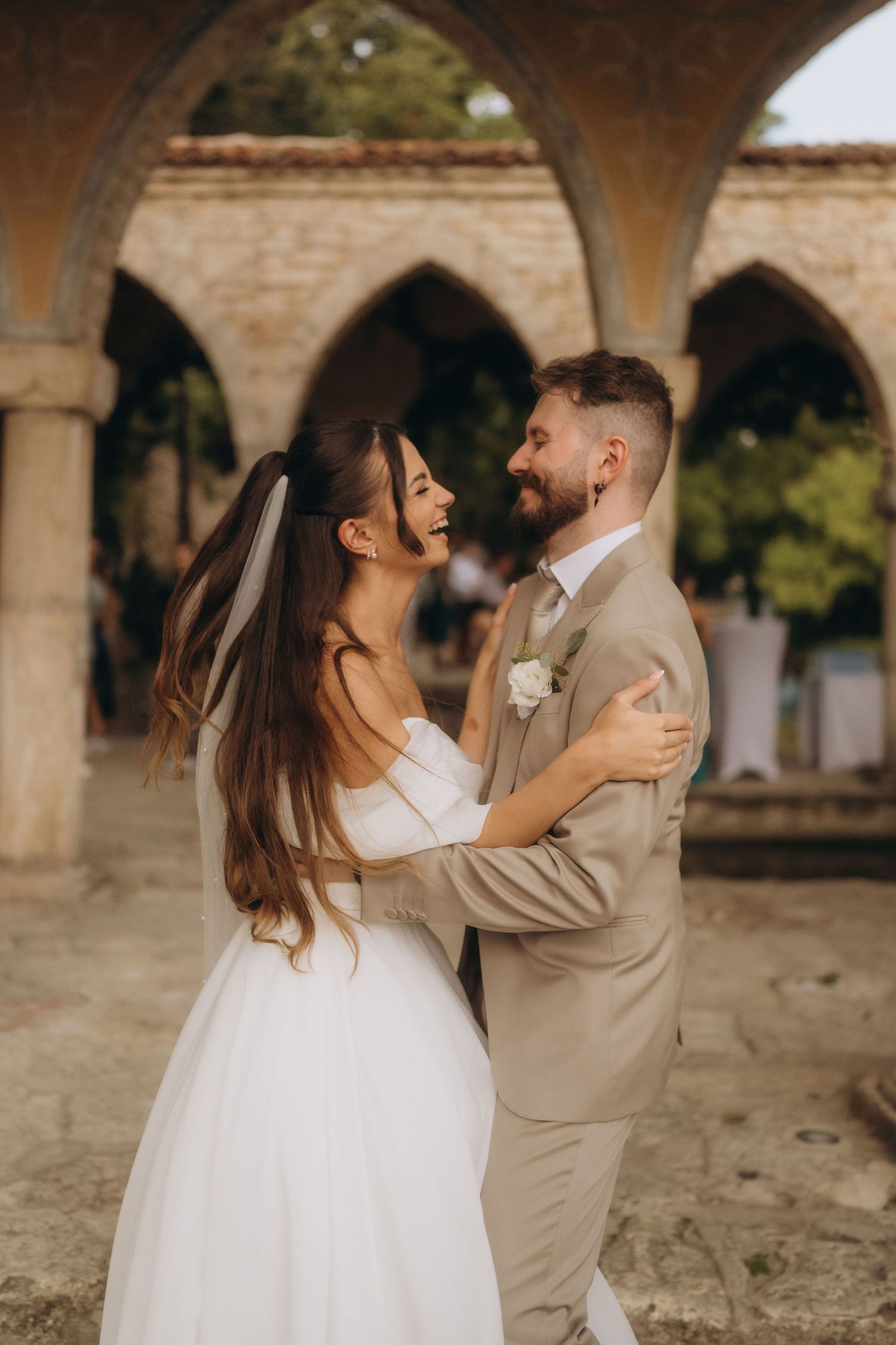 A joyful bride and groom embrace and laugh together outdoors near stone arches at their wedding venue.