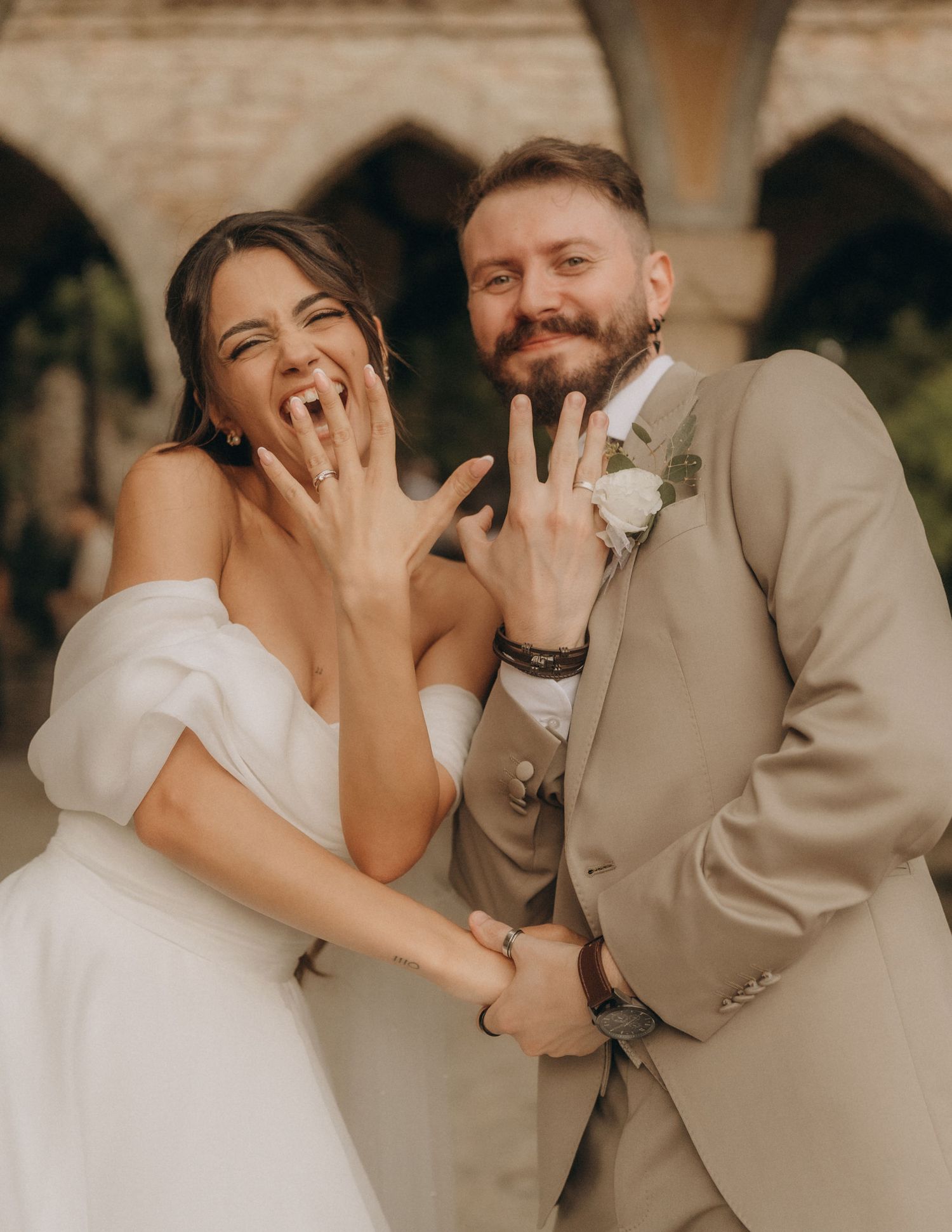 Happy newlyweds showing off wedding rings, bride in white gown and groom in tan suit, celebrating outdoors.