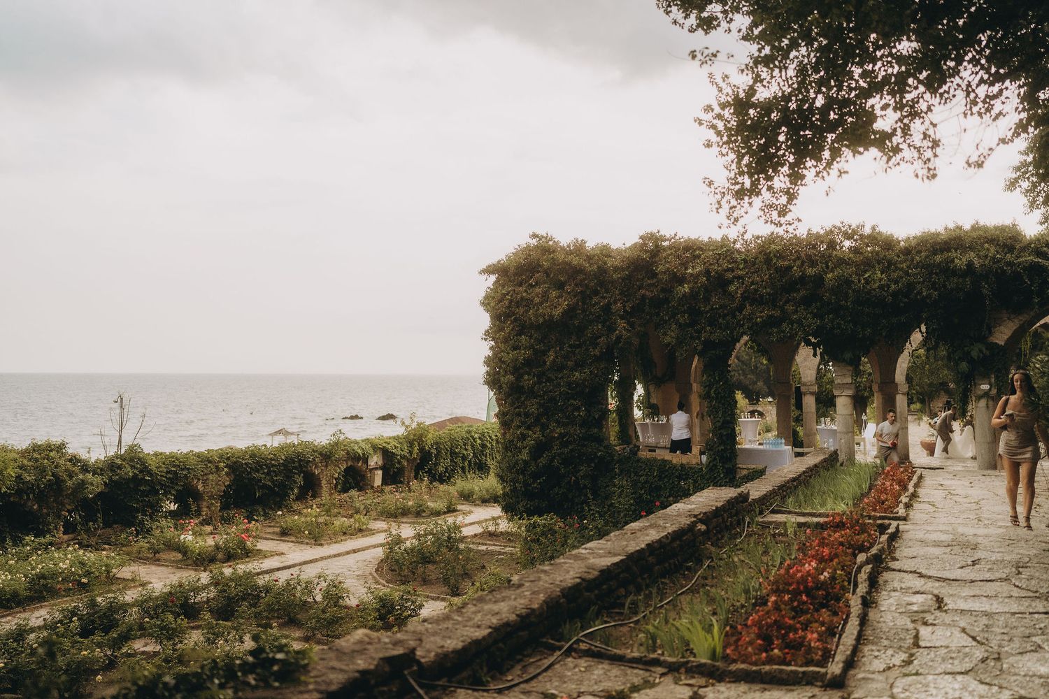 Ivy-covered stone pergola walkway overlooking the sea in a lush coastal garden on a cloudy day.