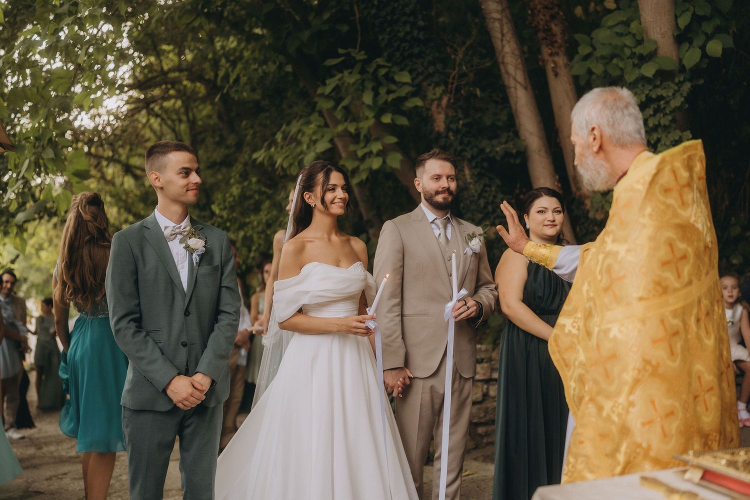 Outdoor wedding ceremony with bride in white dress, groom in tan suit, officiant in gold robes, and guests among green trees.