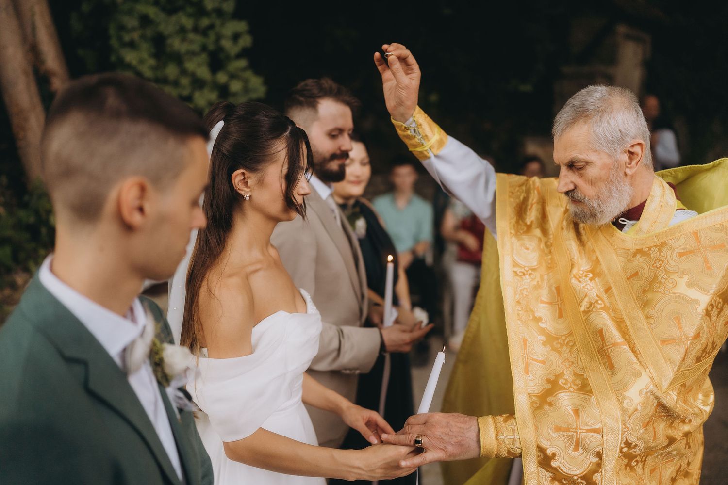 Orthodox priest blessing bride and groom during outdoor wedding ceremony with candles and golden vestments.