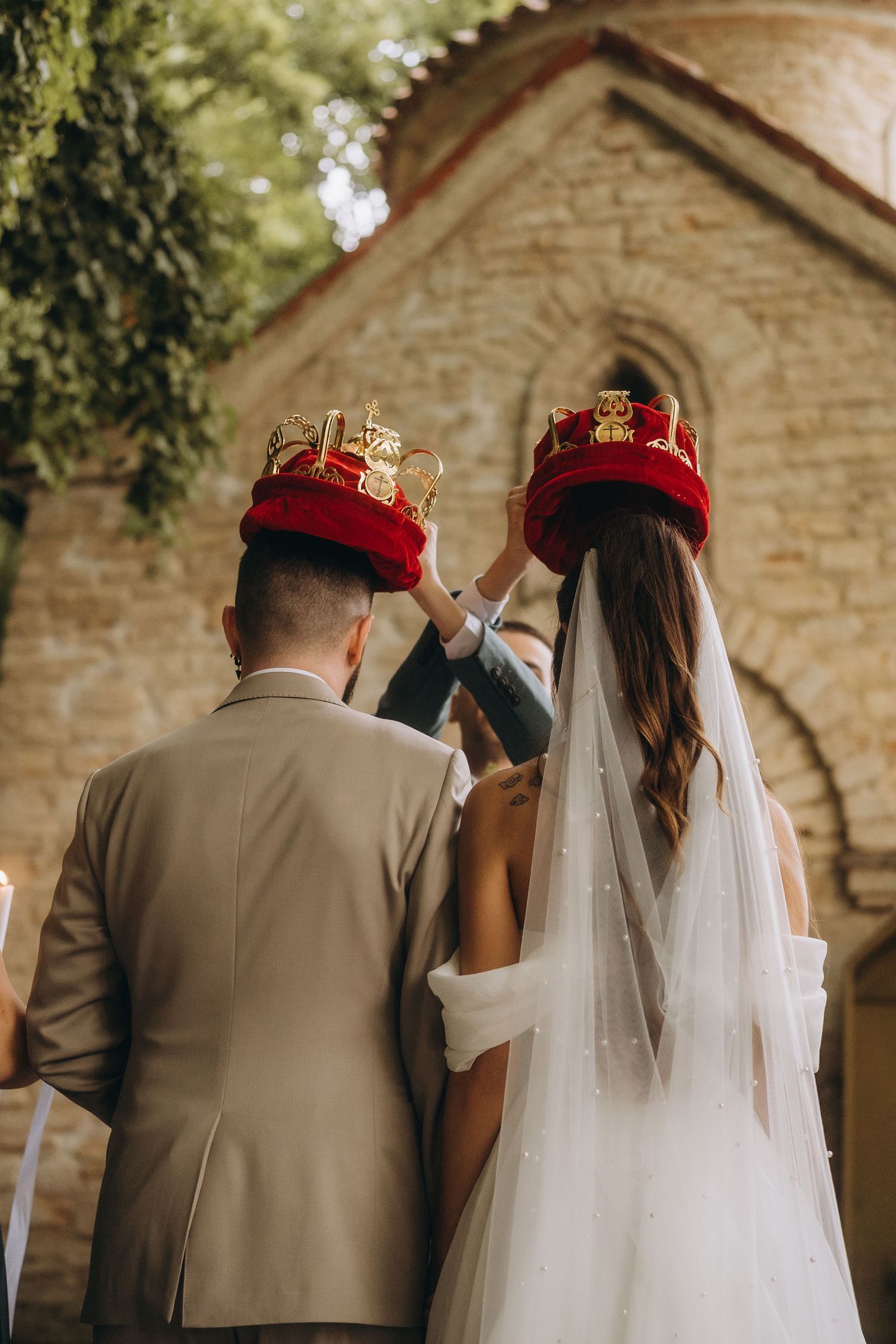 Bride and groom wearing traditional red and gold Orthodox wedding crowns standing before a stone church.