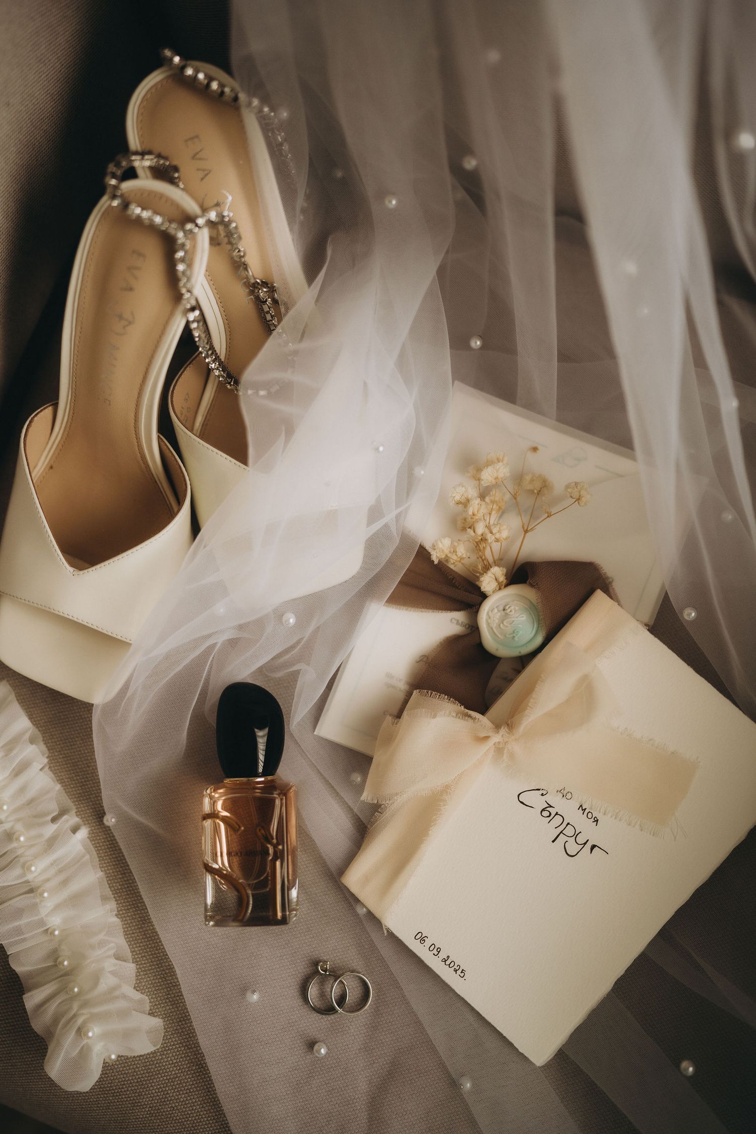 Bridal flat lay with ivory heels, veil, perfume, rings, and wedding invitation on neutral fabric.