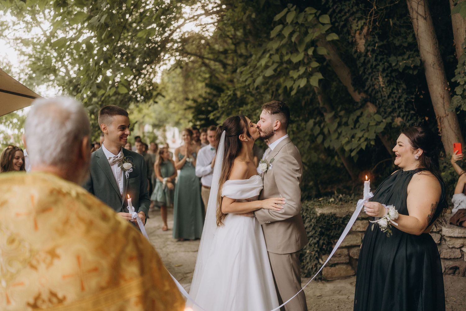 Bride and groom share a kiss during outdoor wedding ceremony surrounded by guests and lush green trees.
