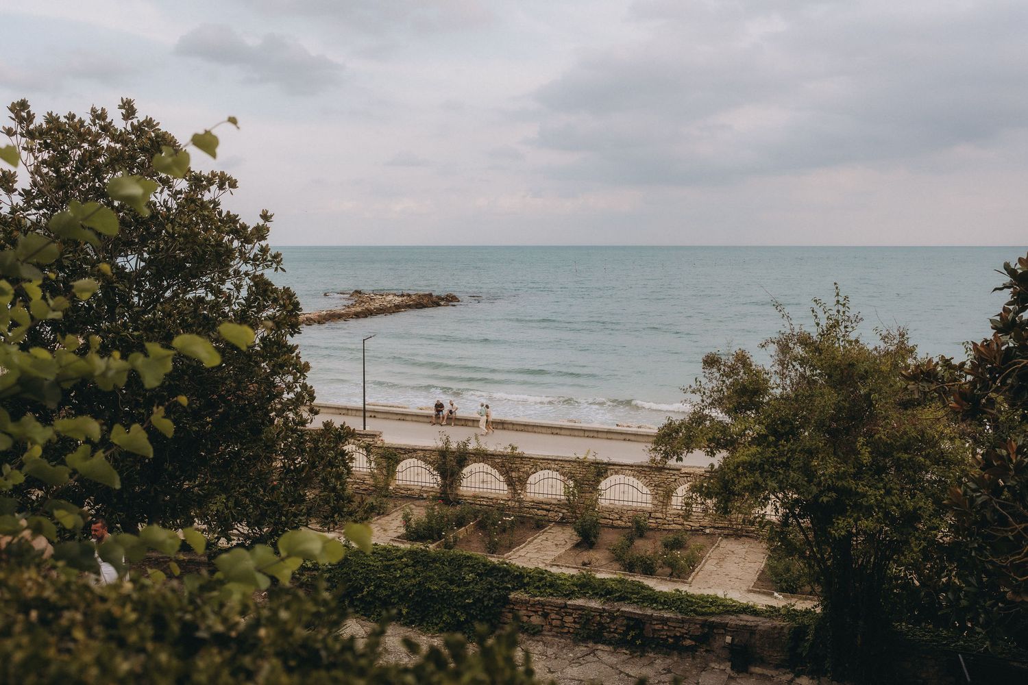 Scenic coastal view with arched promenade, sandy beach, and calm turquoise sea framed by lush green trees.