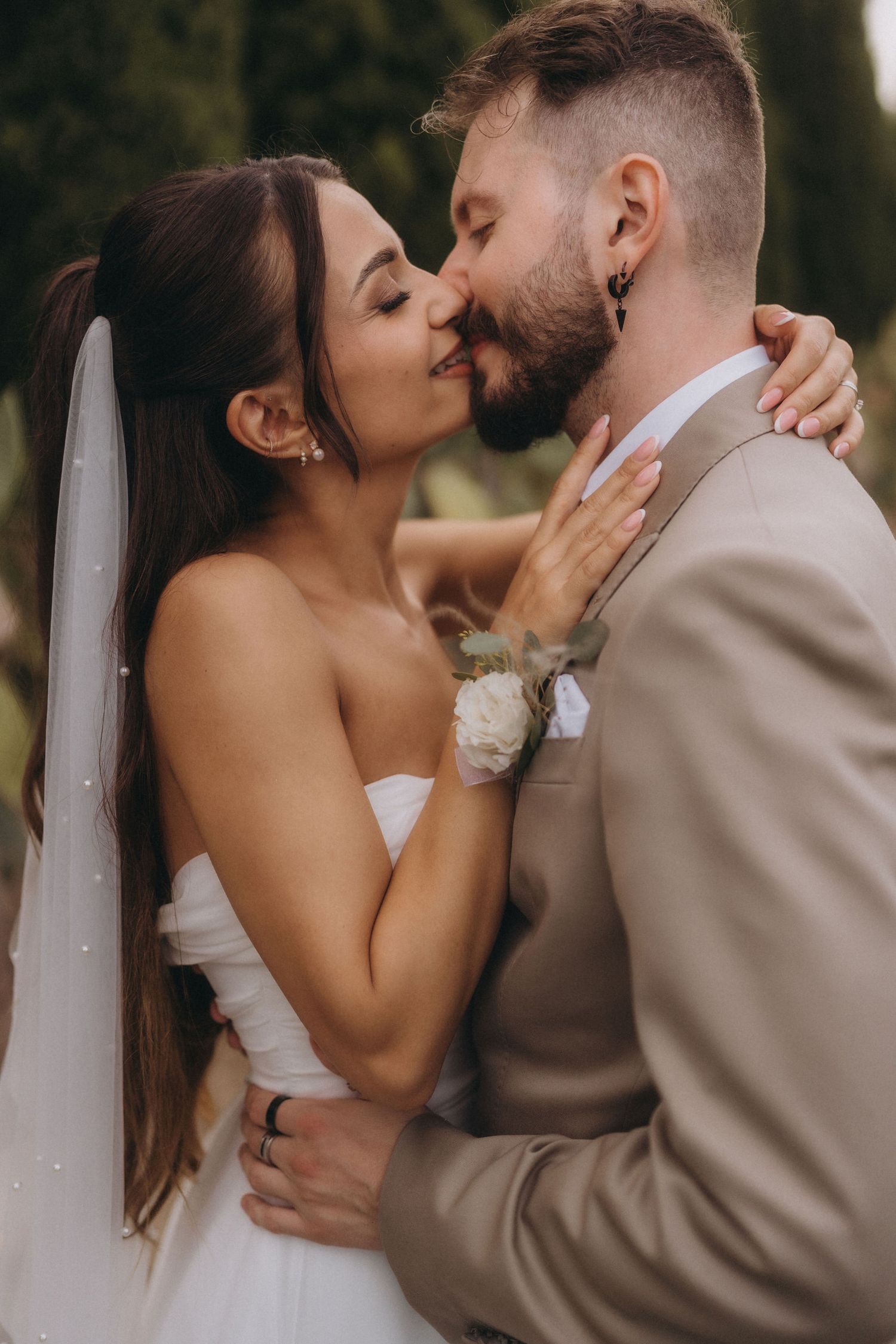 Bride and groom sharing a romantic kiss outdoors, wearing white wedding dress with veil and beige suit with boutonniere.
