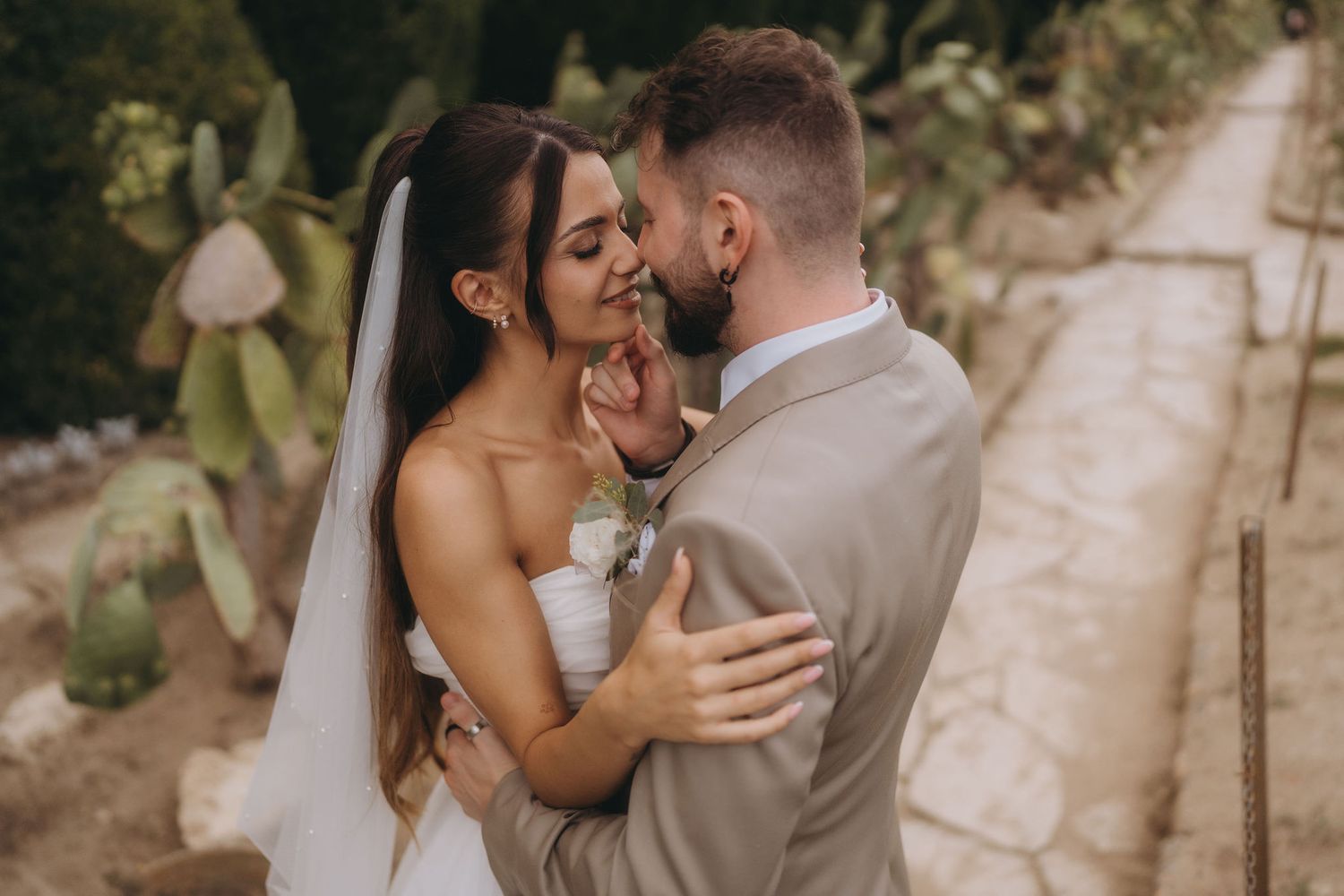 A bride and groom share a romantic moment, nearly kissing outdoors near cactus plants on their wedding day.