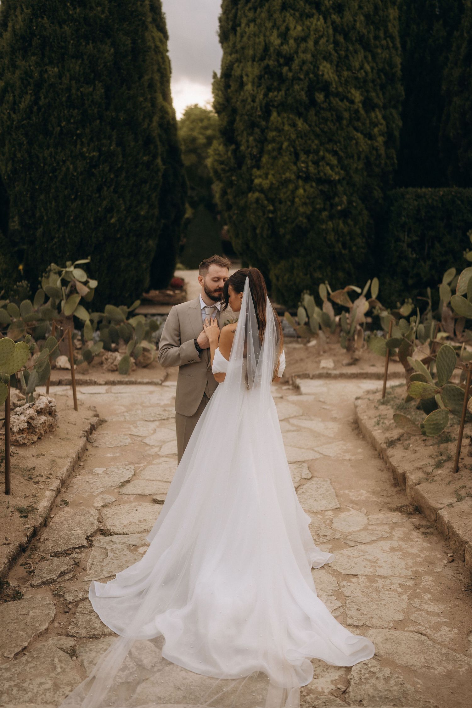 Bride and groom share a romantic moment in a garden pathway lined with cypress trees and cacti.