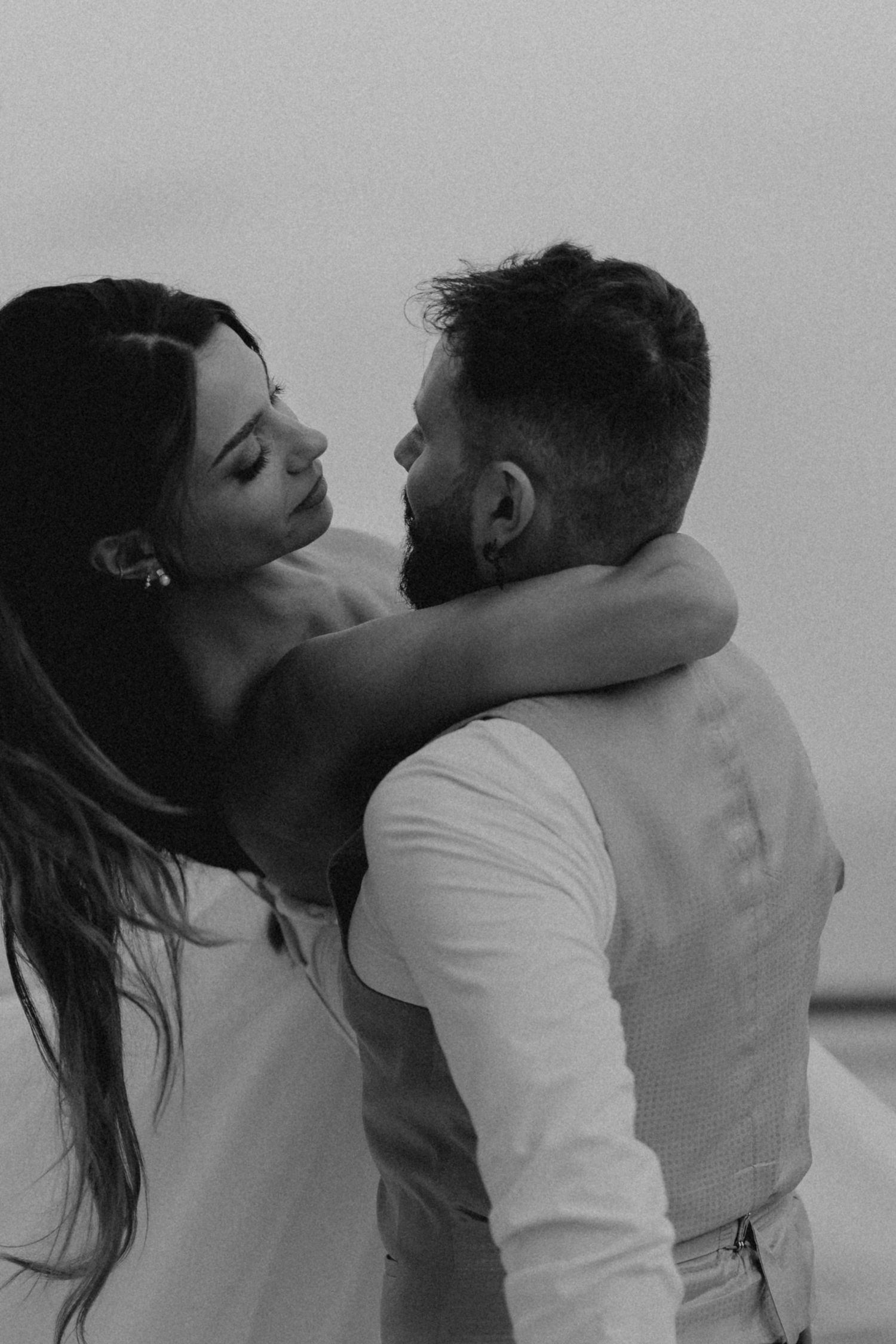 Black and white photo of a couple embracing and gazing at each other, dressed in white, romantic outdoor setting.
