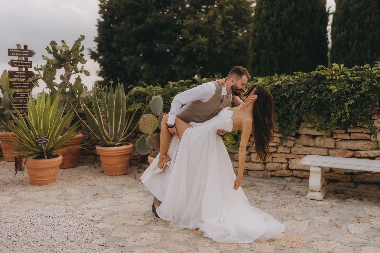 Groom dips bride for a romantic kiss in a rustic garden with cacti and stone wall backdrop.