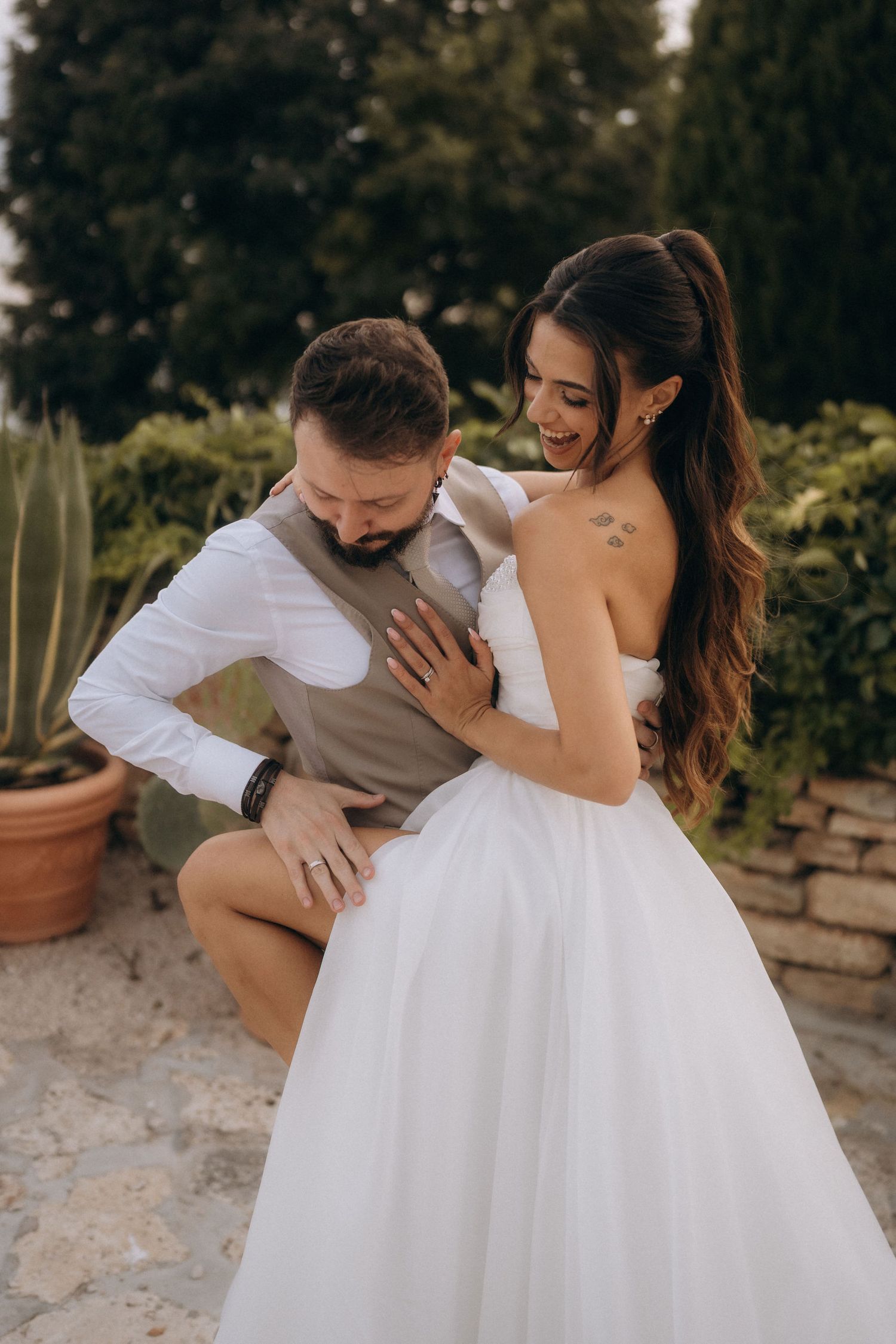 Bride in white strapless gown laughing as groom in vest playfully dips during outdoor wedding photo session.