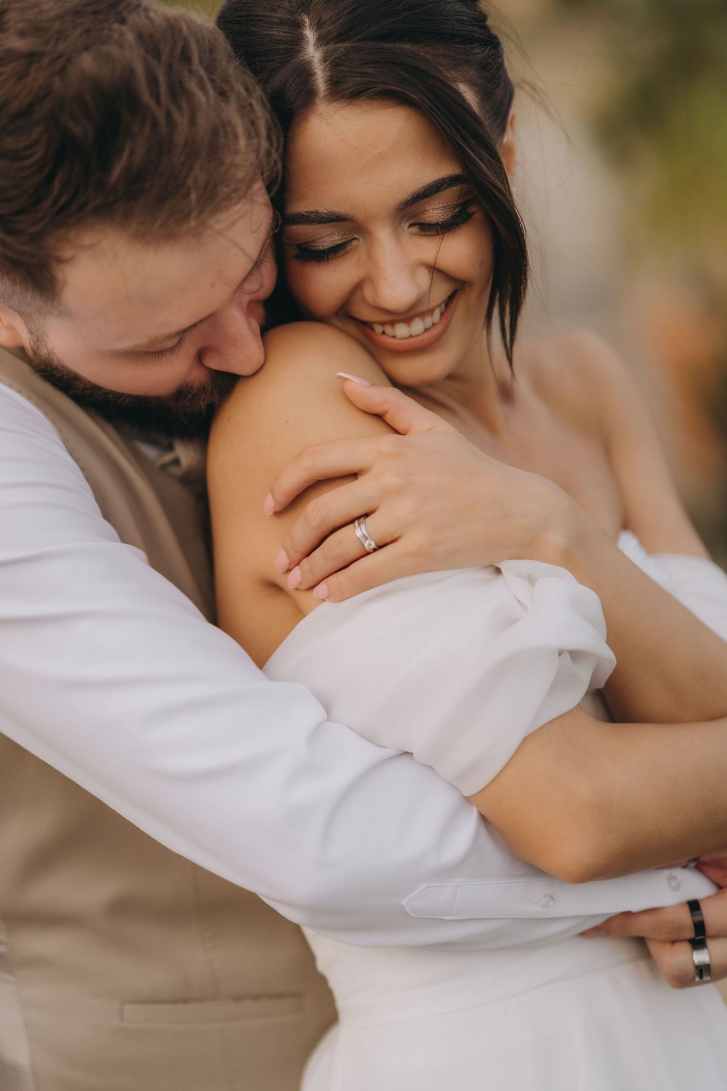 Smiling bride with diamond ring embraces groom in white shirt during romantic outdoor wedding portrait.