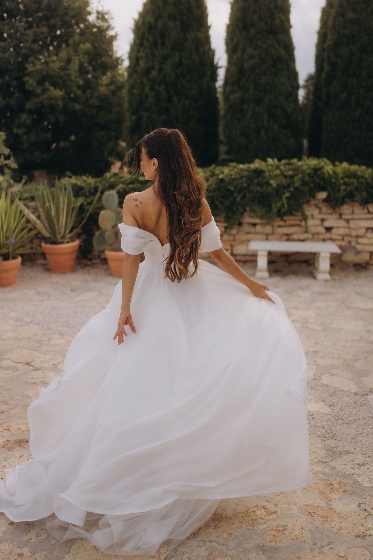 Bride in off-shoulder white ballgown twirling in garden with cypress trees and stone wall backdrop.