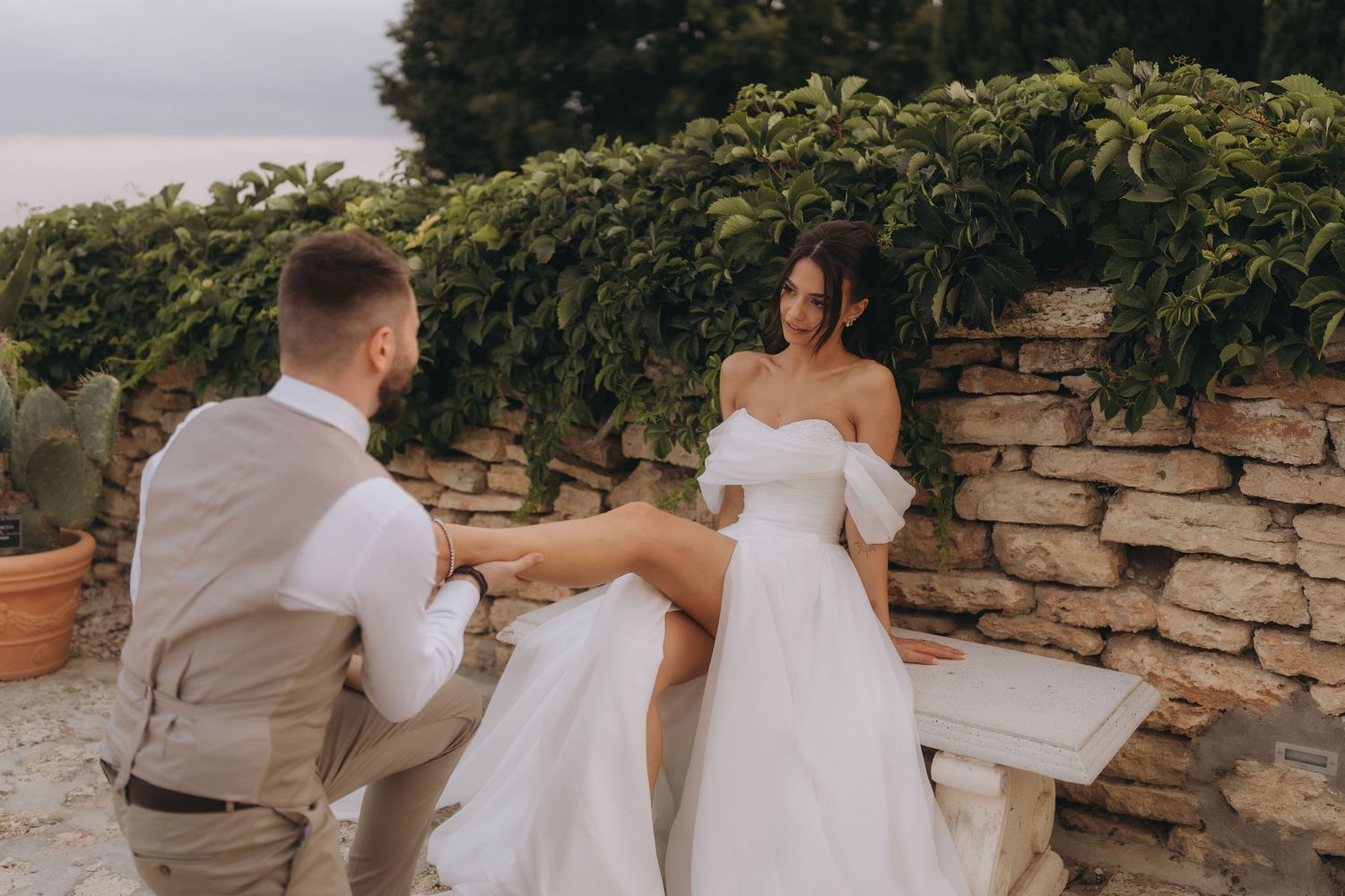Bride in white off-shoulder gown sits on stone bench as groom kneels before her in lush garden setting.