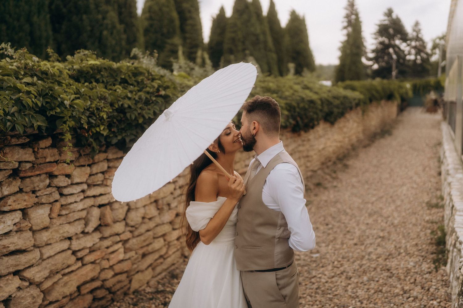 Bride and groom share a kiss behind white parasol along stone wall in elegant outdoor wedding setting.