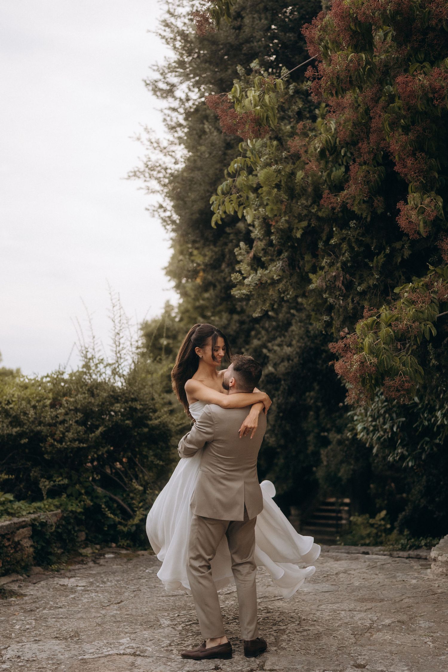 Groom lifting bride in white dress, embracing outdoors among lush green and red foliage on their wedding day.