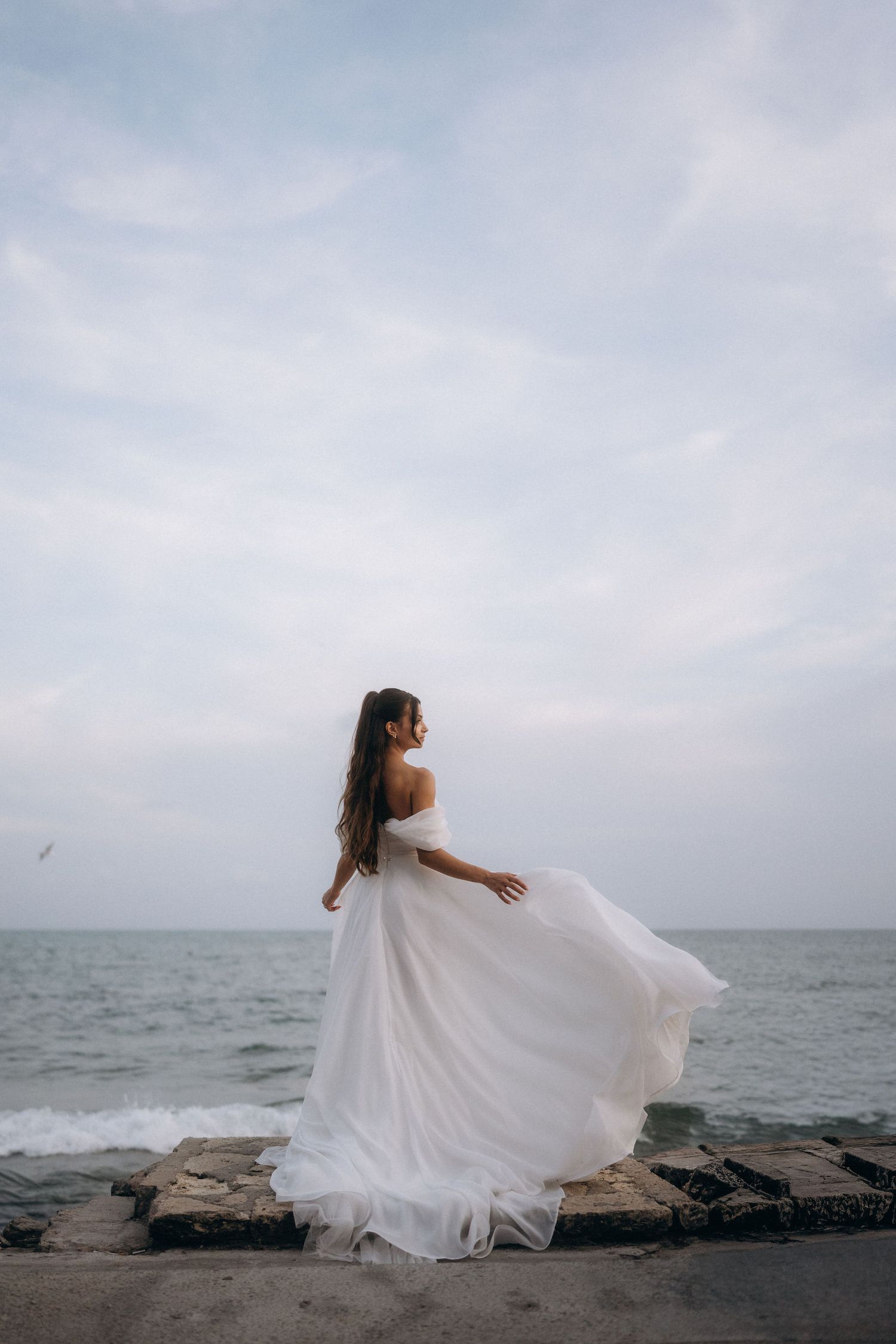 A bride in a flowing white off-shoulder gown stands on a seaside pier, her dress billowing in the wind.