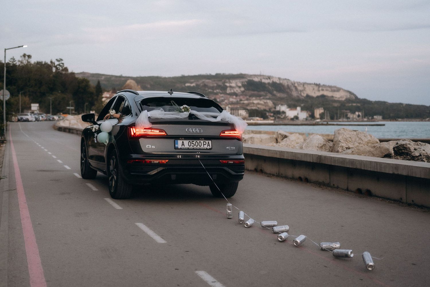 Black Audi SUV parked on coastal road with scattered cans trailing behind it near seaside cliffs.