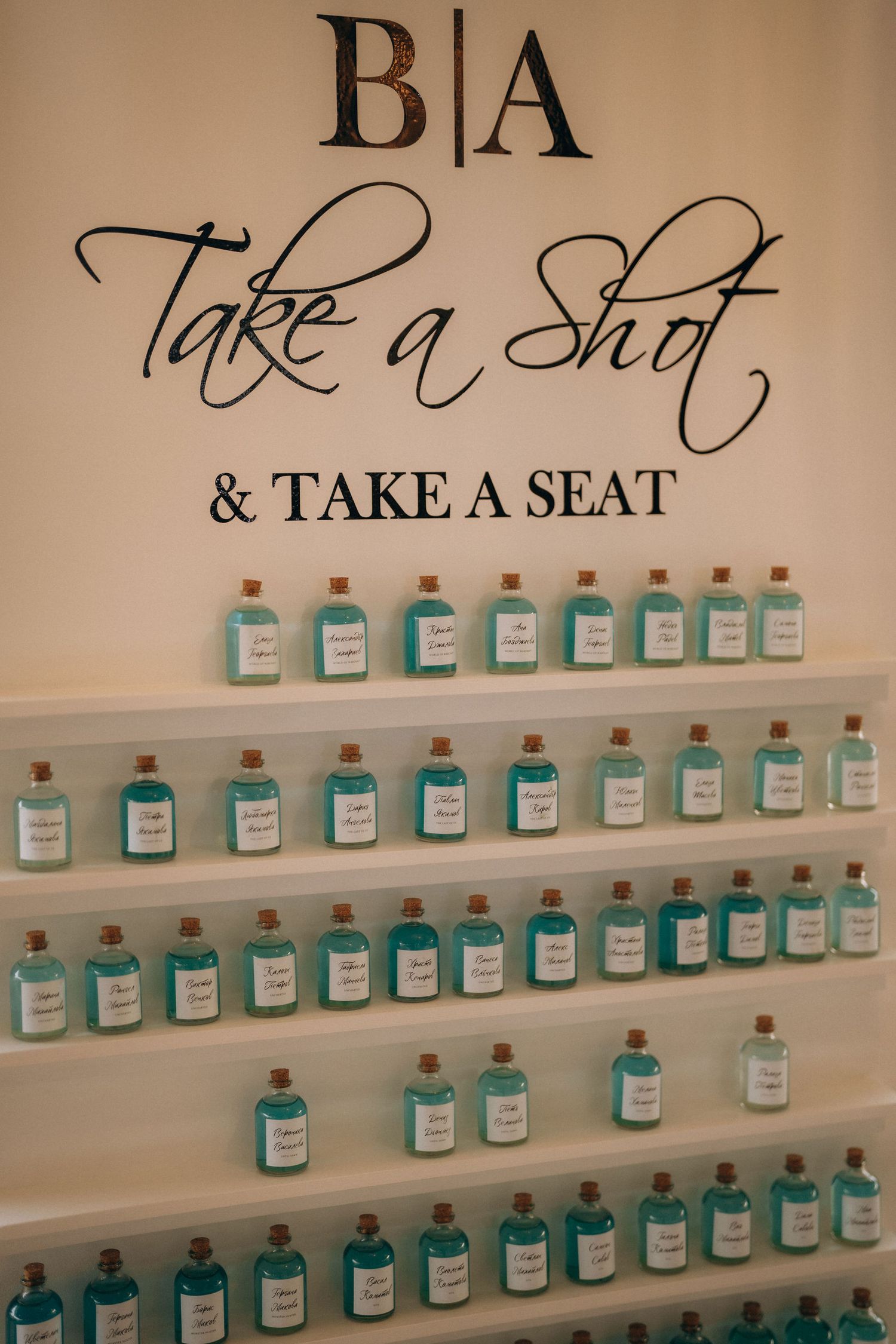 Wedding seating chart display with teal shot bottles on white shelves, reading Take a Shot & Take a Seat.