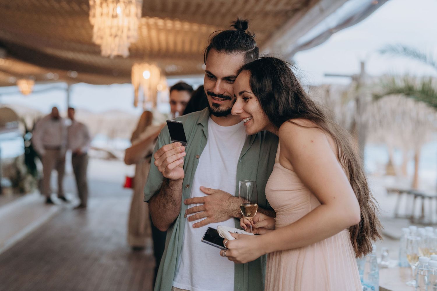 Couple smiling at a smartphone while holding wine glasses at an elegant outdoor beach party event.