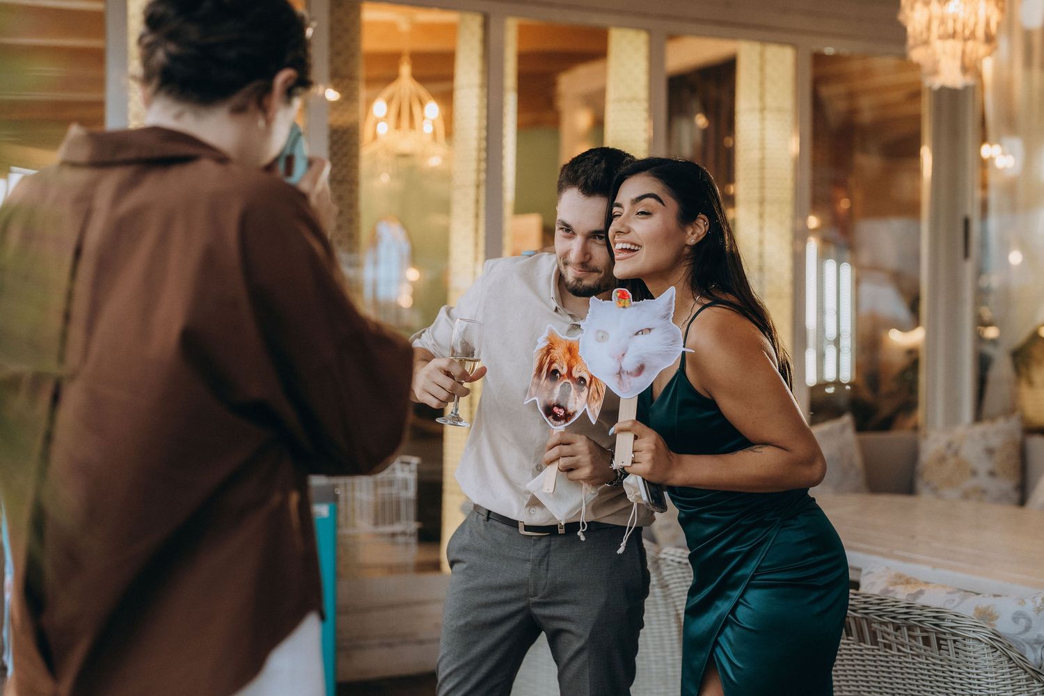 Couple posing together at a party, woman in green dress embracing man holding wine glass while photographer takes their photo.